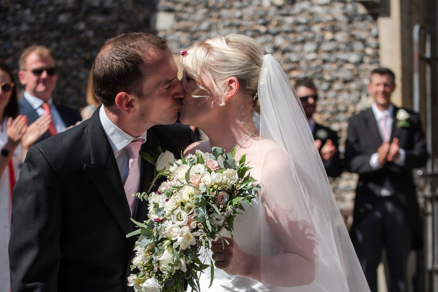 Bride and groom sharing a romantic kiss outside a stone church surrounded by applauding guests.  St Michaels church, bray
