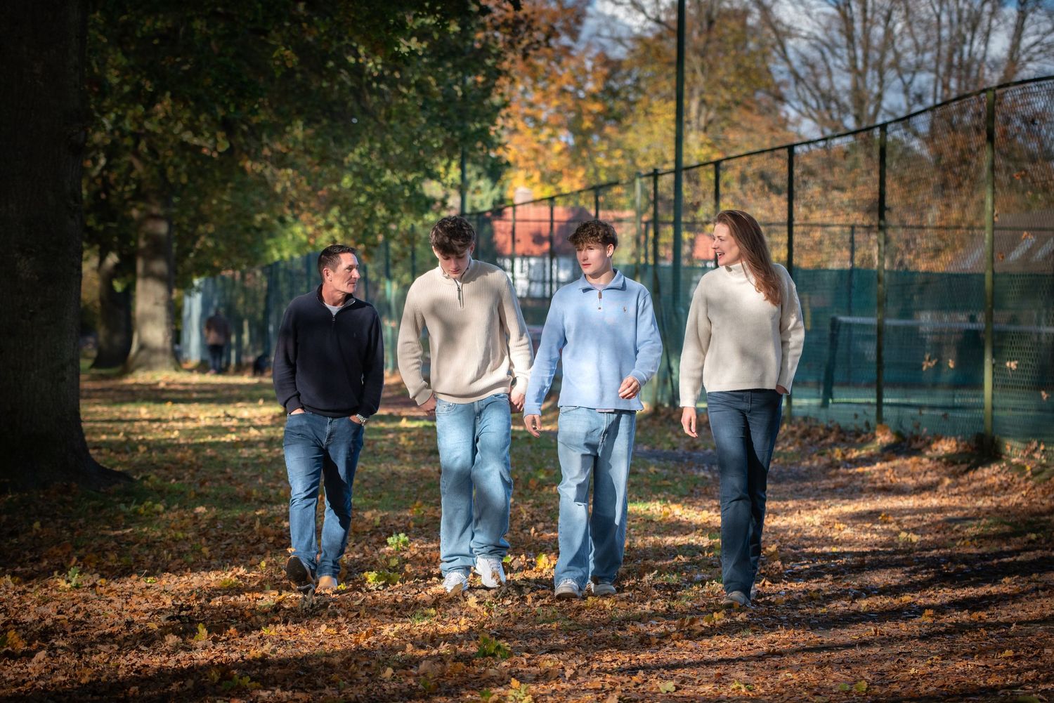 Family of four walking together along a path in an autumn park near a green fence.