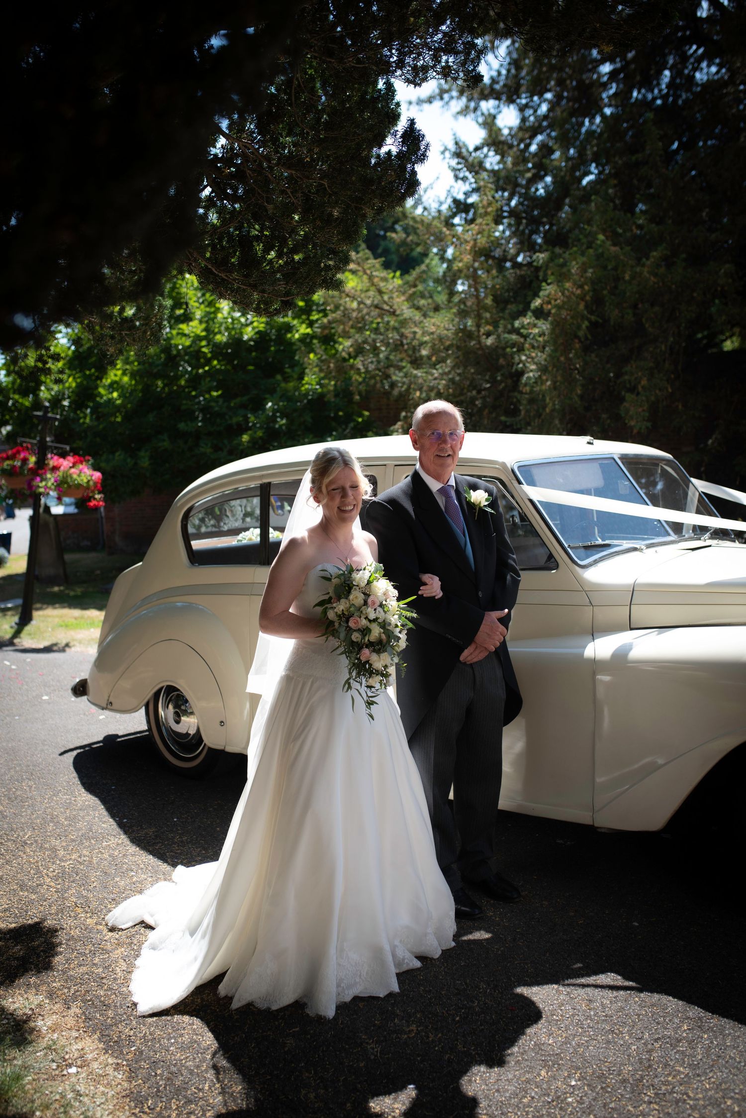 Bride in white gown with bouquet and father in dark suit posing beside a vintage cream wedding car. St Michaels Church Bray