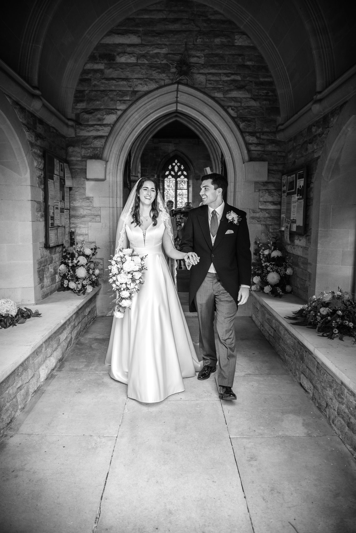 Black and white photo of a bride and groom walking hand in hand through a stone church archway. St Saviours Church Brockenhurst