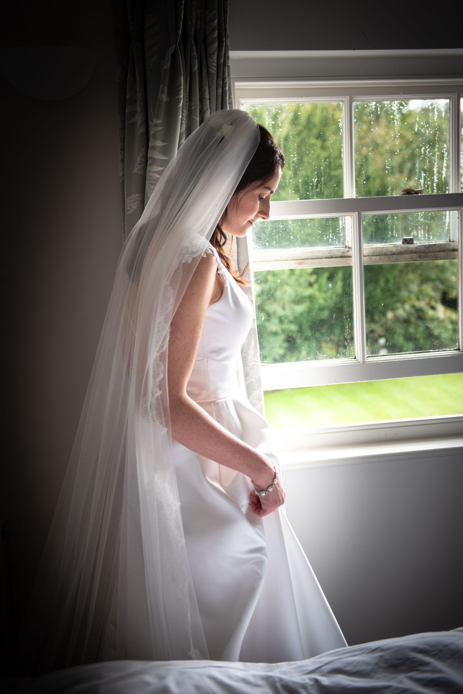 Bride in white gown and veil standing by a window holding her bouquet.