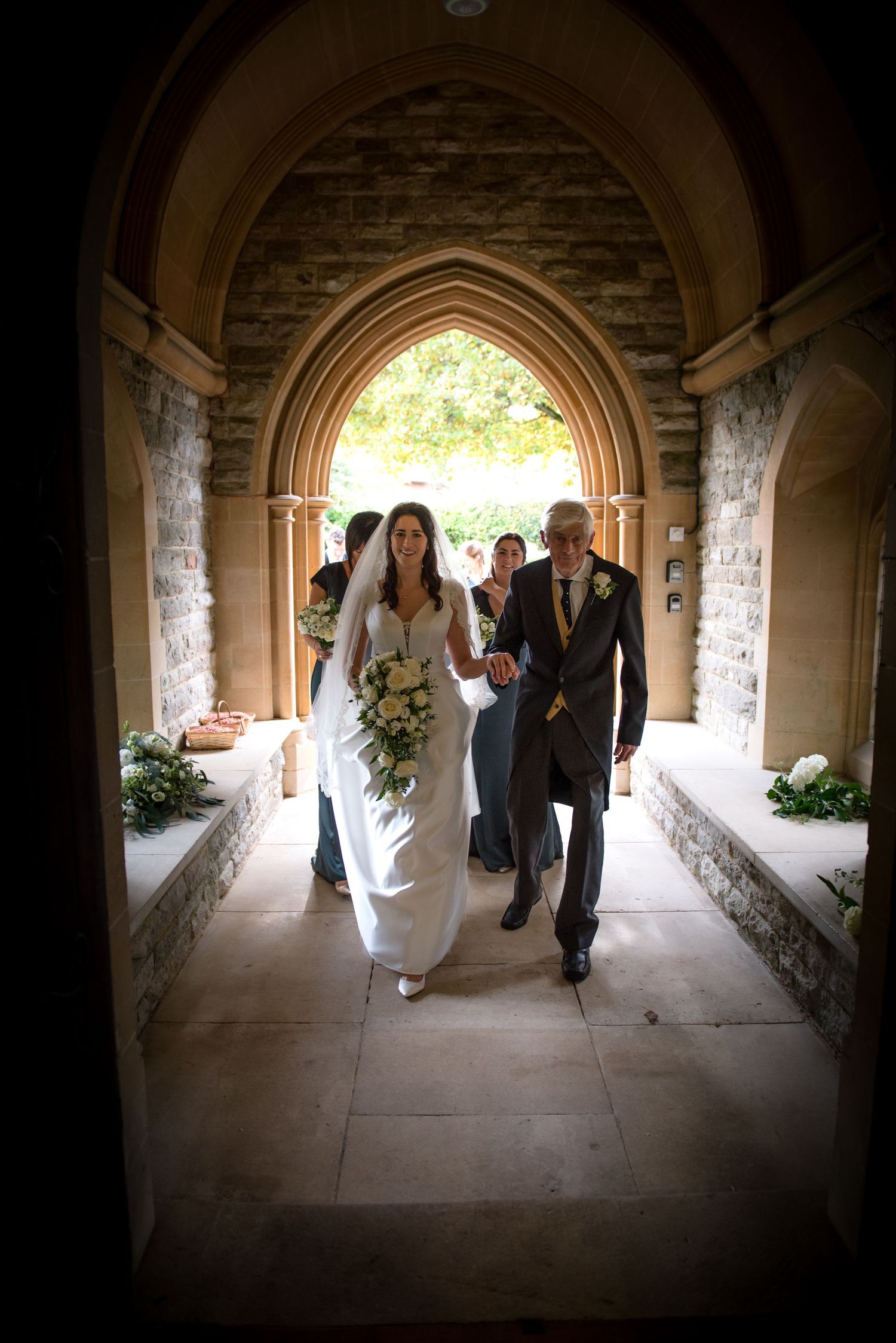 Bride walks down a stone church archway corridor with her father before the wedding ceremony.