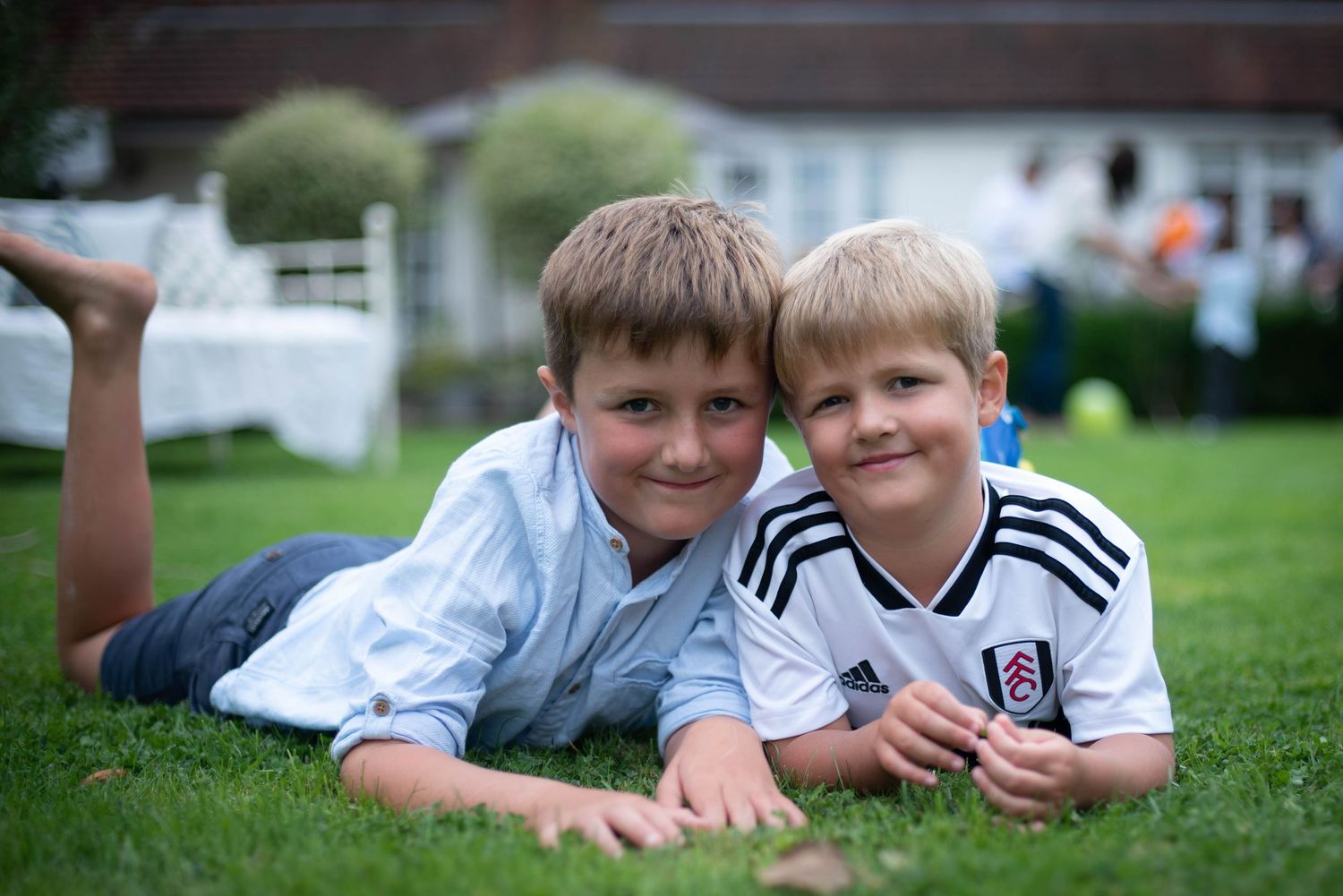 Two young boys in white shirts lying on green grass smiling at camera.