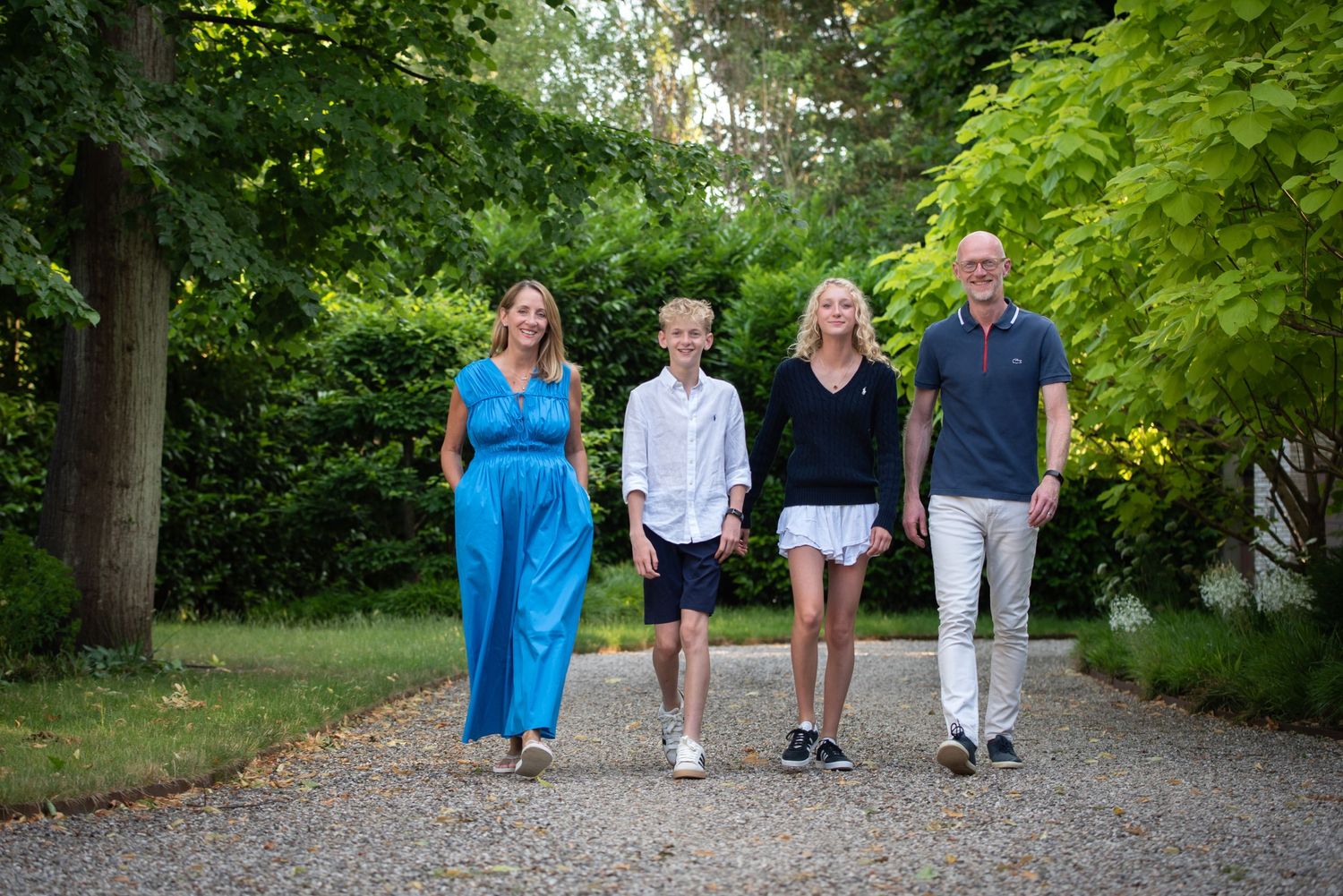 A family of four walks together along a tree-lined gravel path in summer.