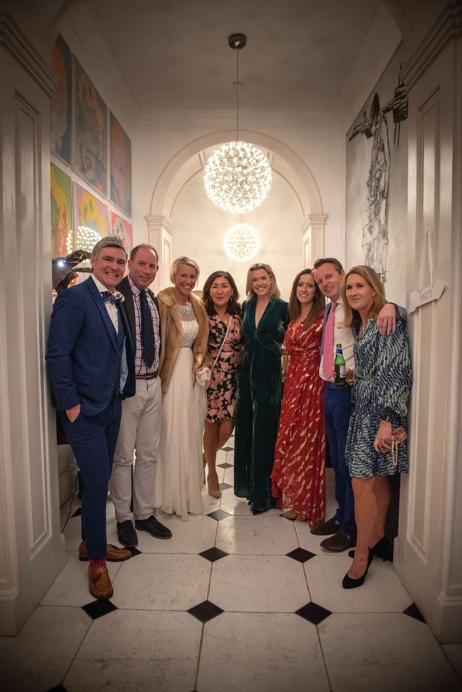 Group of eight wedding guests posing together in an elegant white hallway with arched ceiling. Wedding reception
