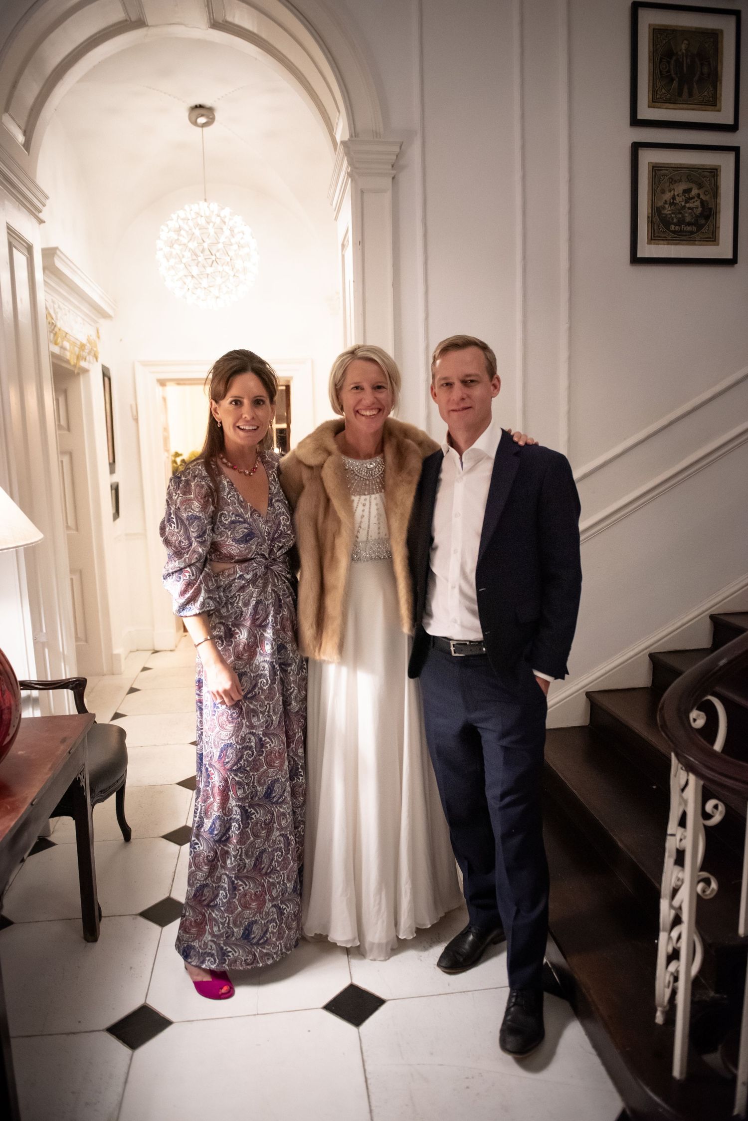 Bride in white gown with fur stole poses with two guests in a grand white hallway.