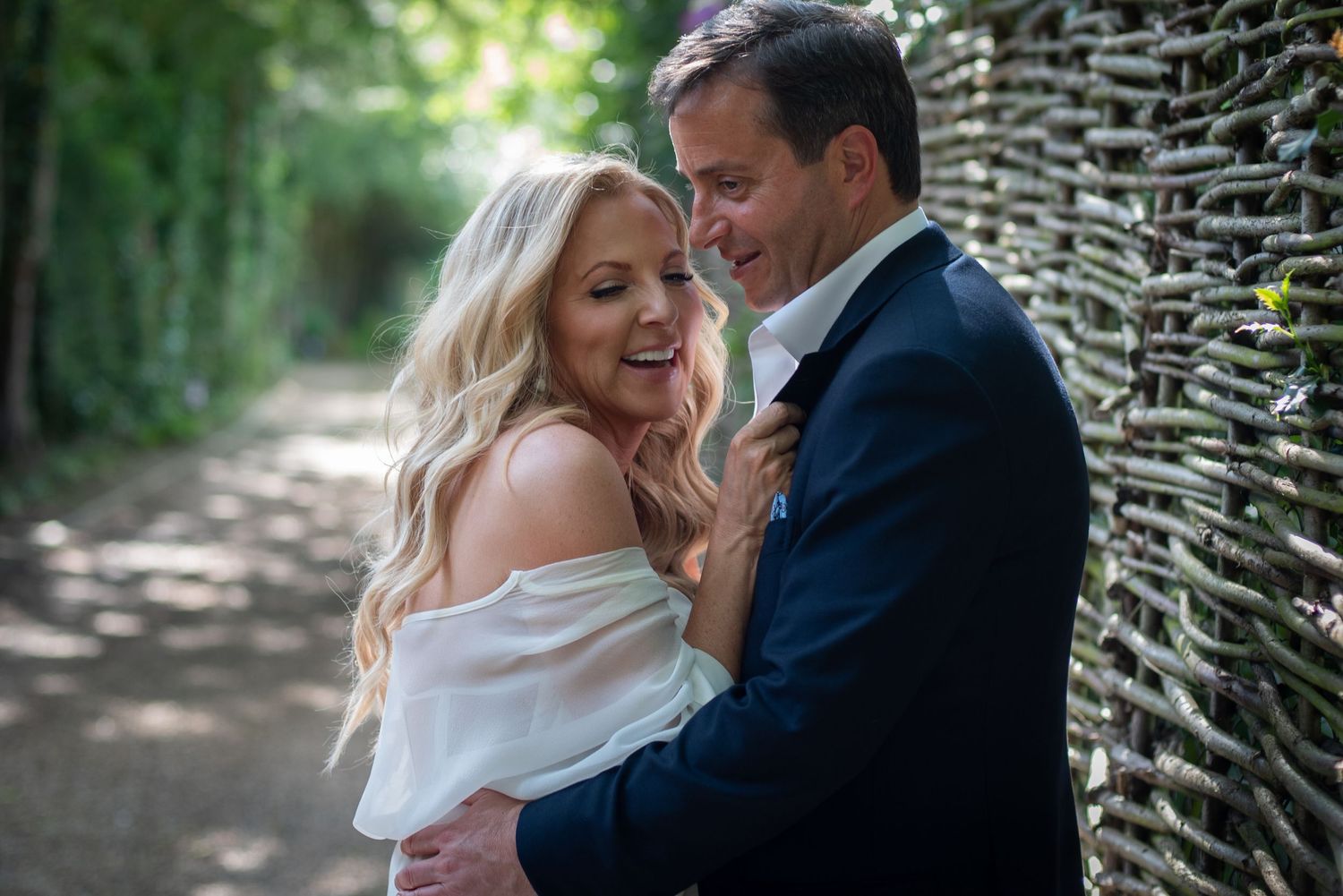 Laughing bride and groom embracing on a tree-lined path in soft natural light.