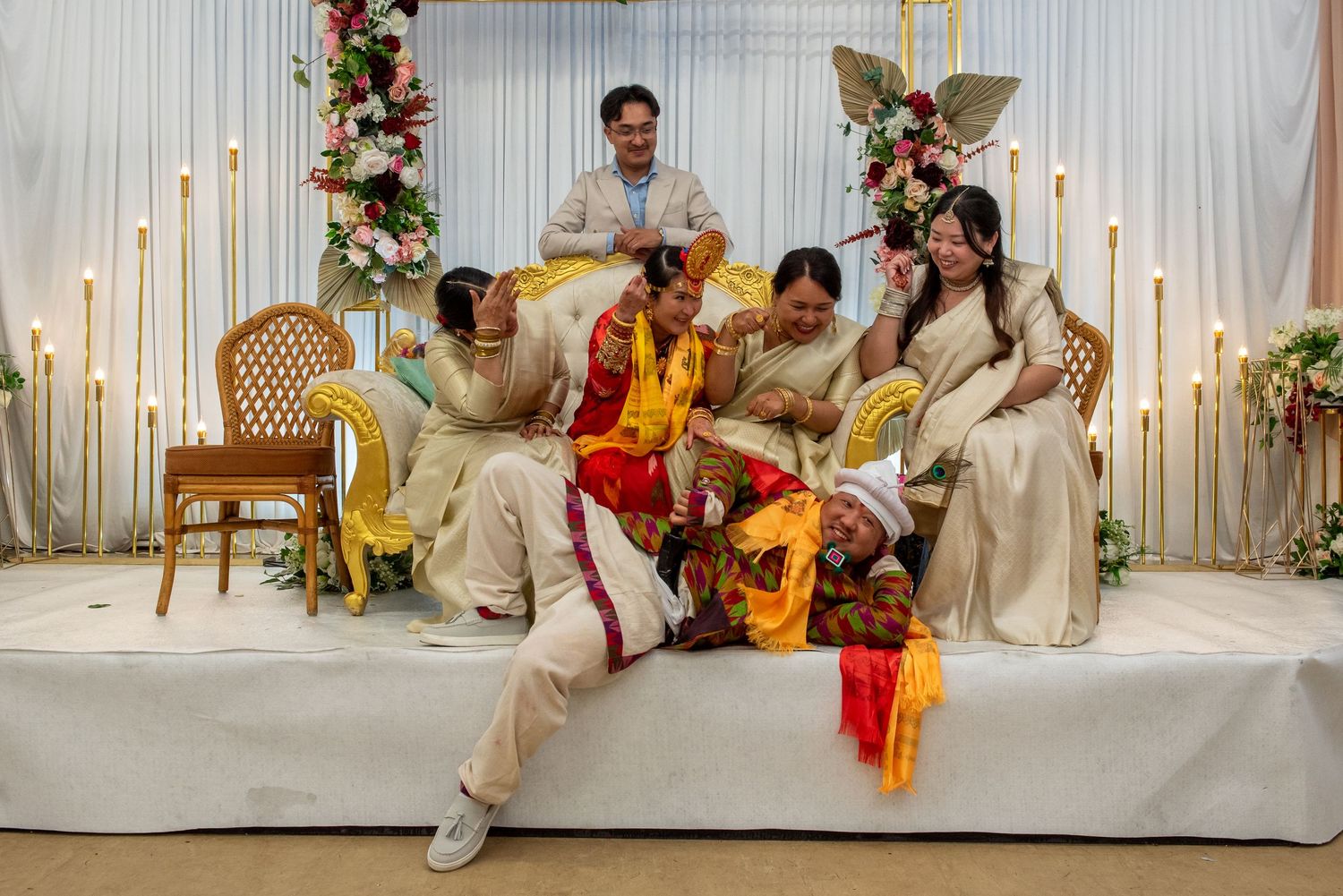 Joyful wedding party in colorful traditional attire posing together on a floral decorated stage.