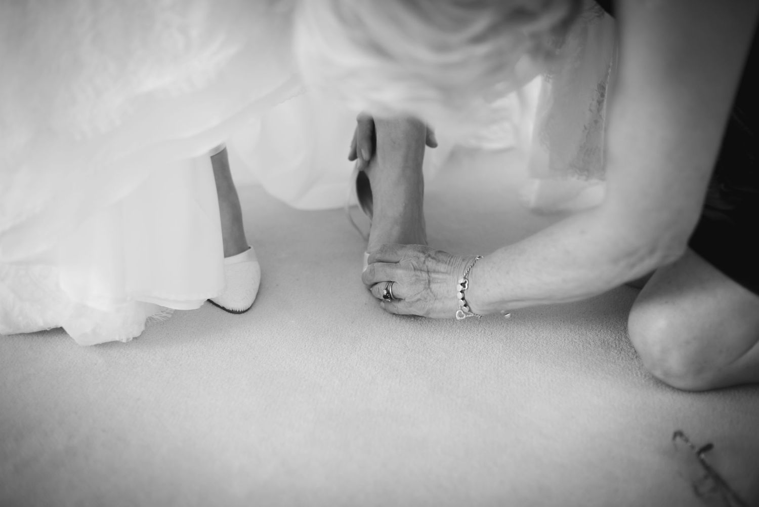 Black and white image of bride putting on heeled wedding shoes while seated on the floor.