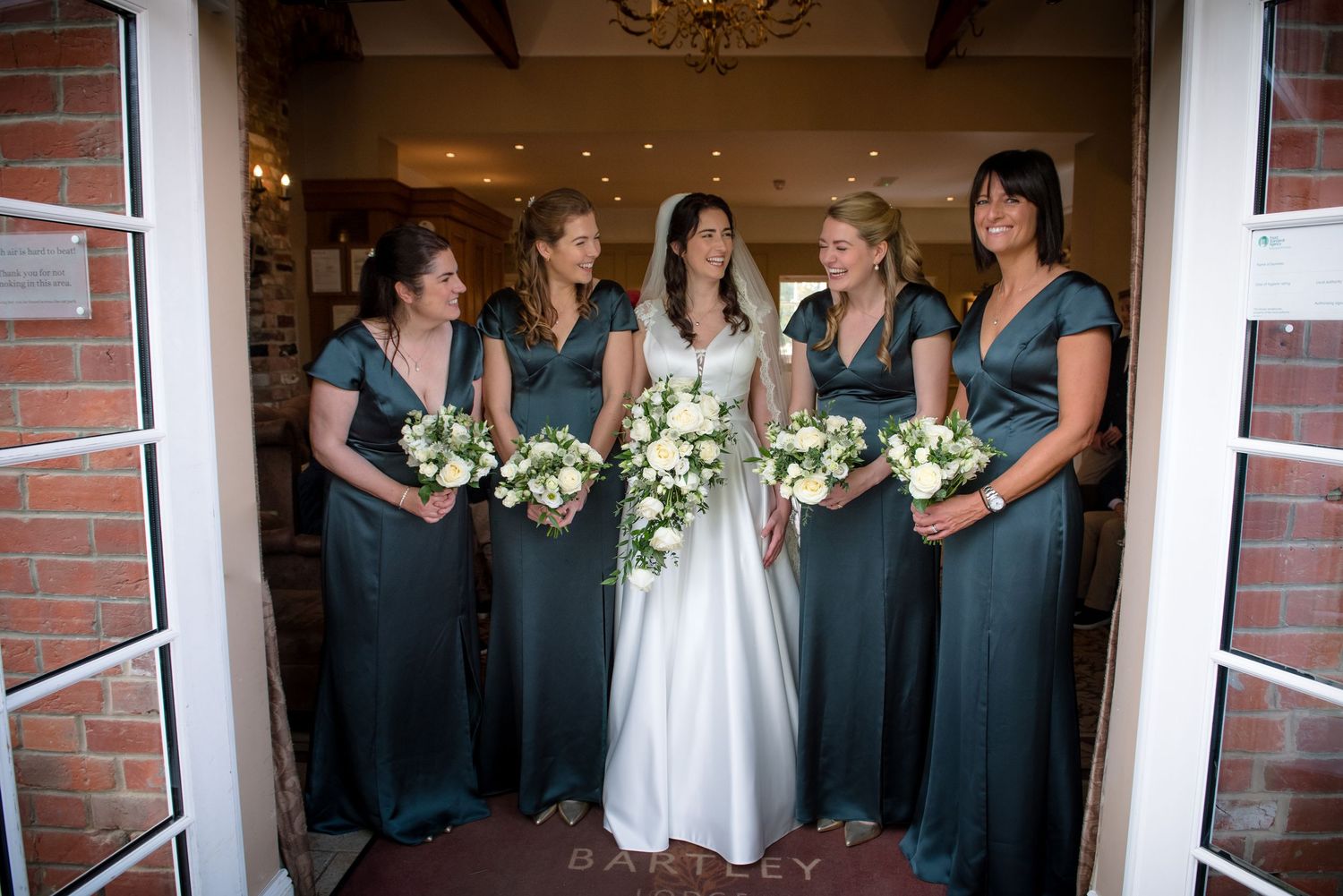 Bride with four bridesmaids in teal dresses holding white bouquets at venue.