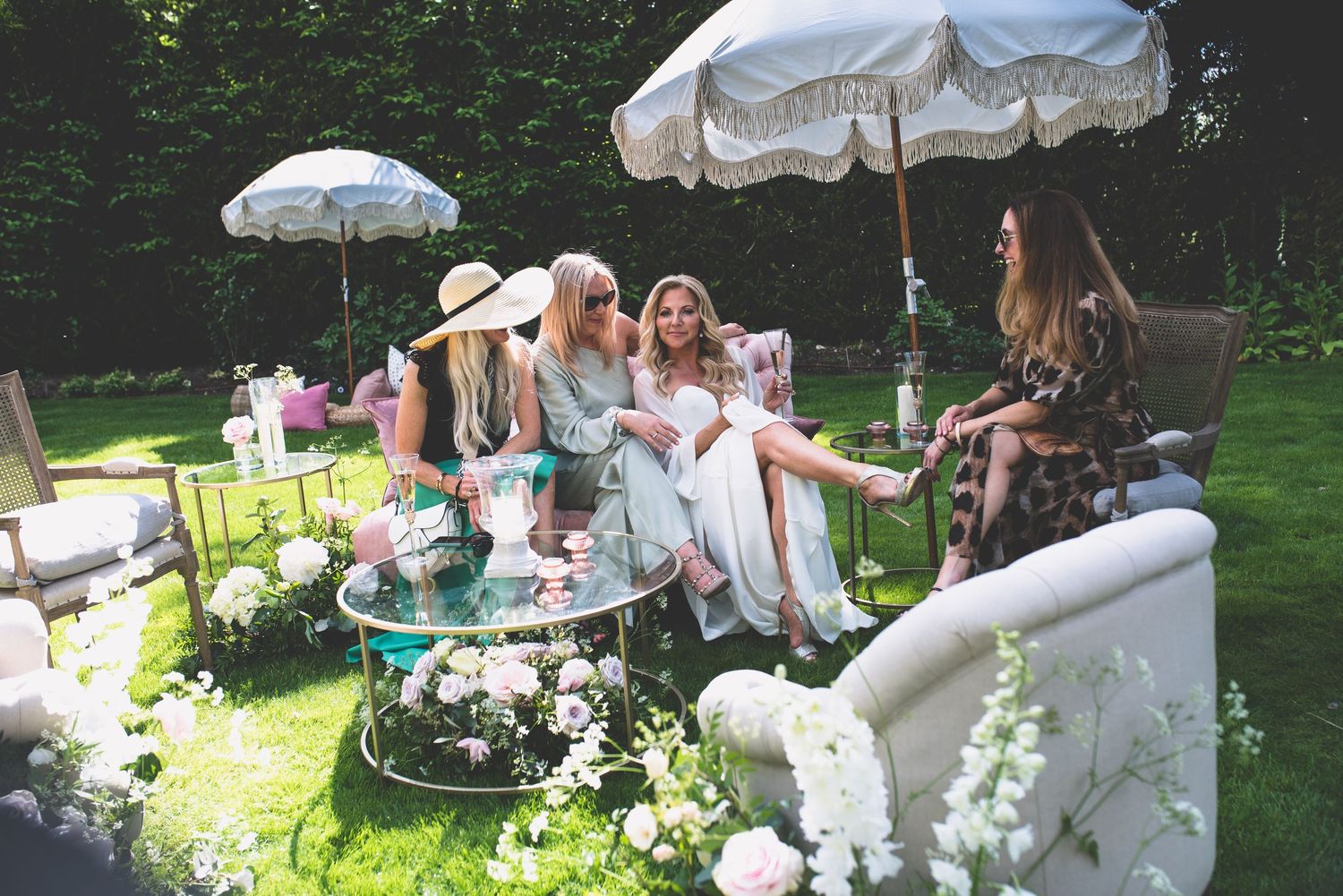 Four women socializing at a stylish garden party seated under white parasols surrounded by floral arrangements.