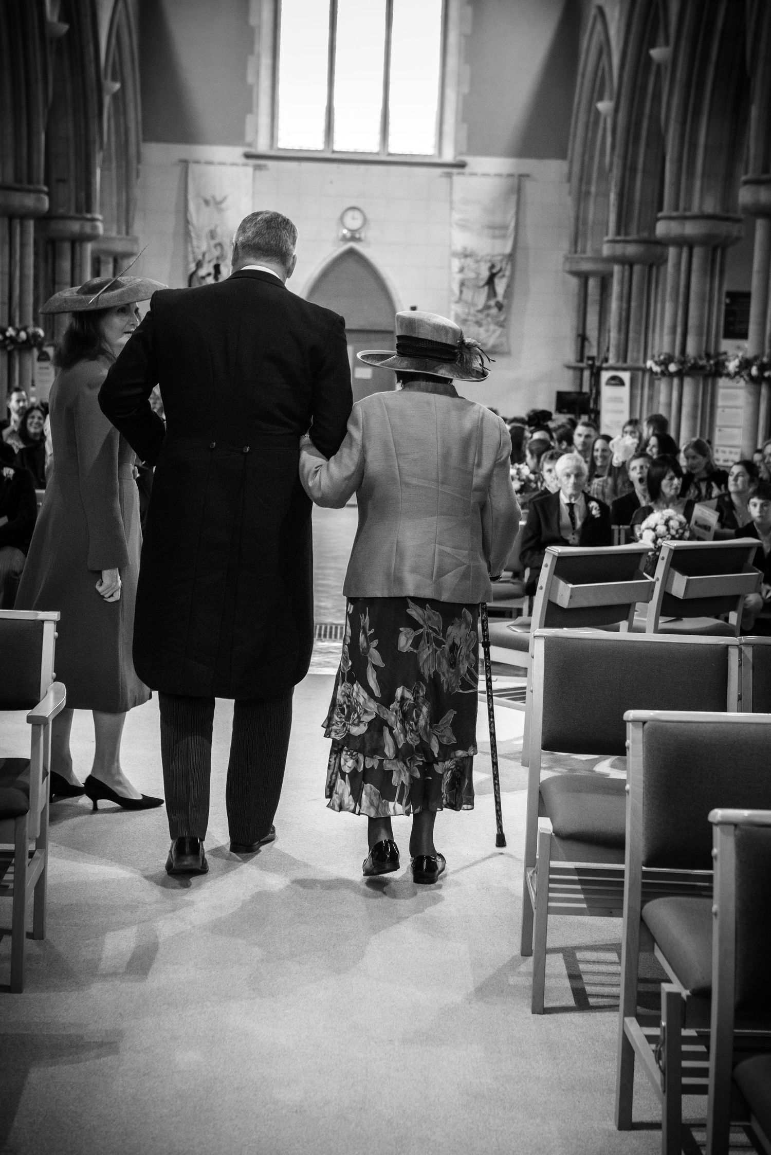 Black and white photo of elderly woman with cane being escorted down church aisle by gentleman.
