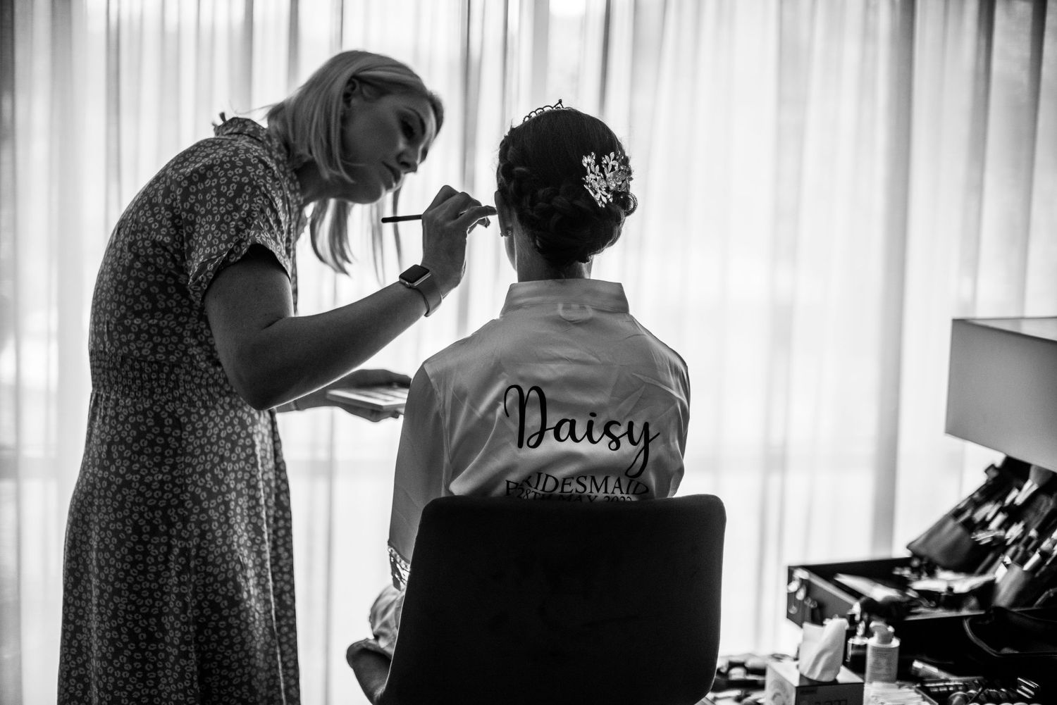 Makeup artist applying makeup to bride Daisy in a personalized robe during wedding preparations.