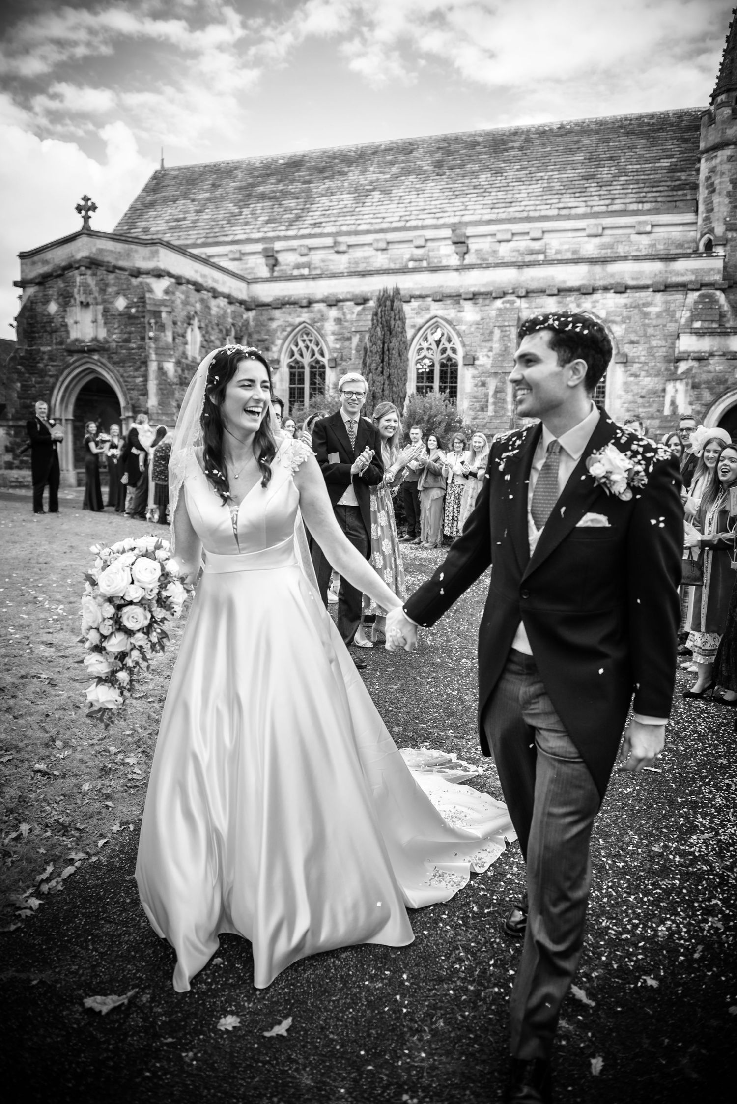 A joyful bride and groom walk hand in hand outside a historic stone church after their wedding ceremony.