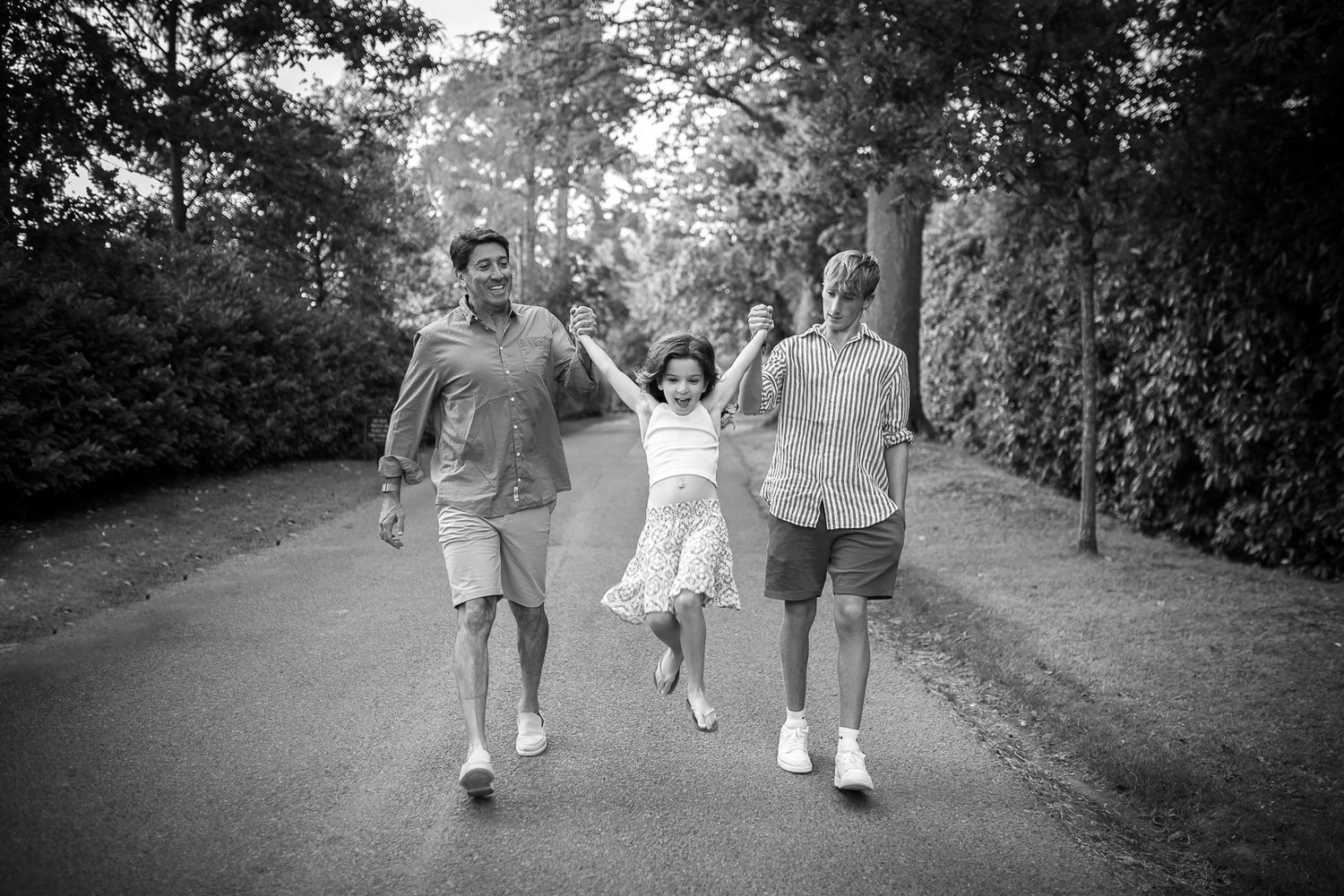 Black and white photo of a happy family walking on a tree-lined path, parents swinging their young daughter.