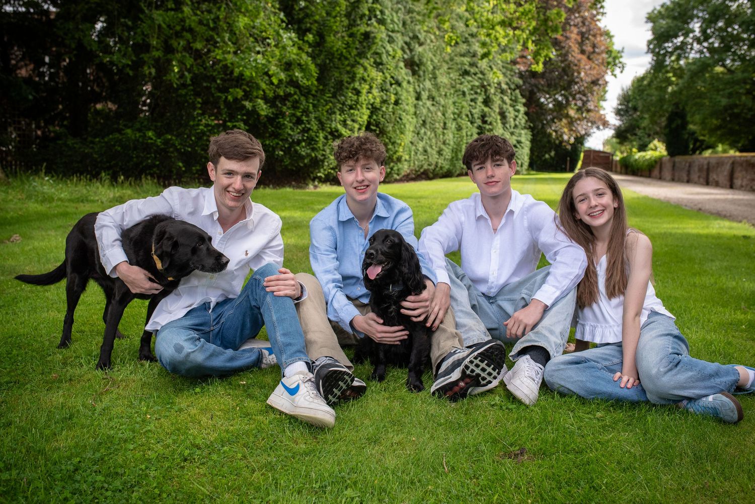 Four teenagers sitting on grass with two dogs in a vibrant green garden on a sunny day.