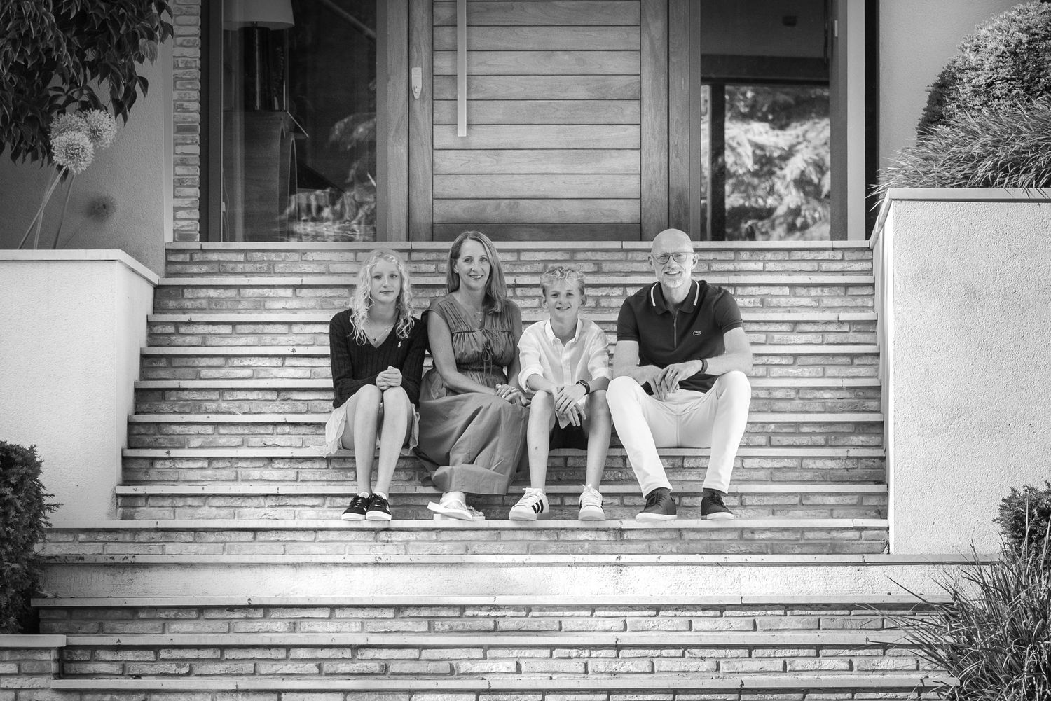 Family of four sitting on stone steps in black and white portrait outside elegant home entrance.