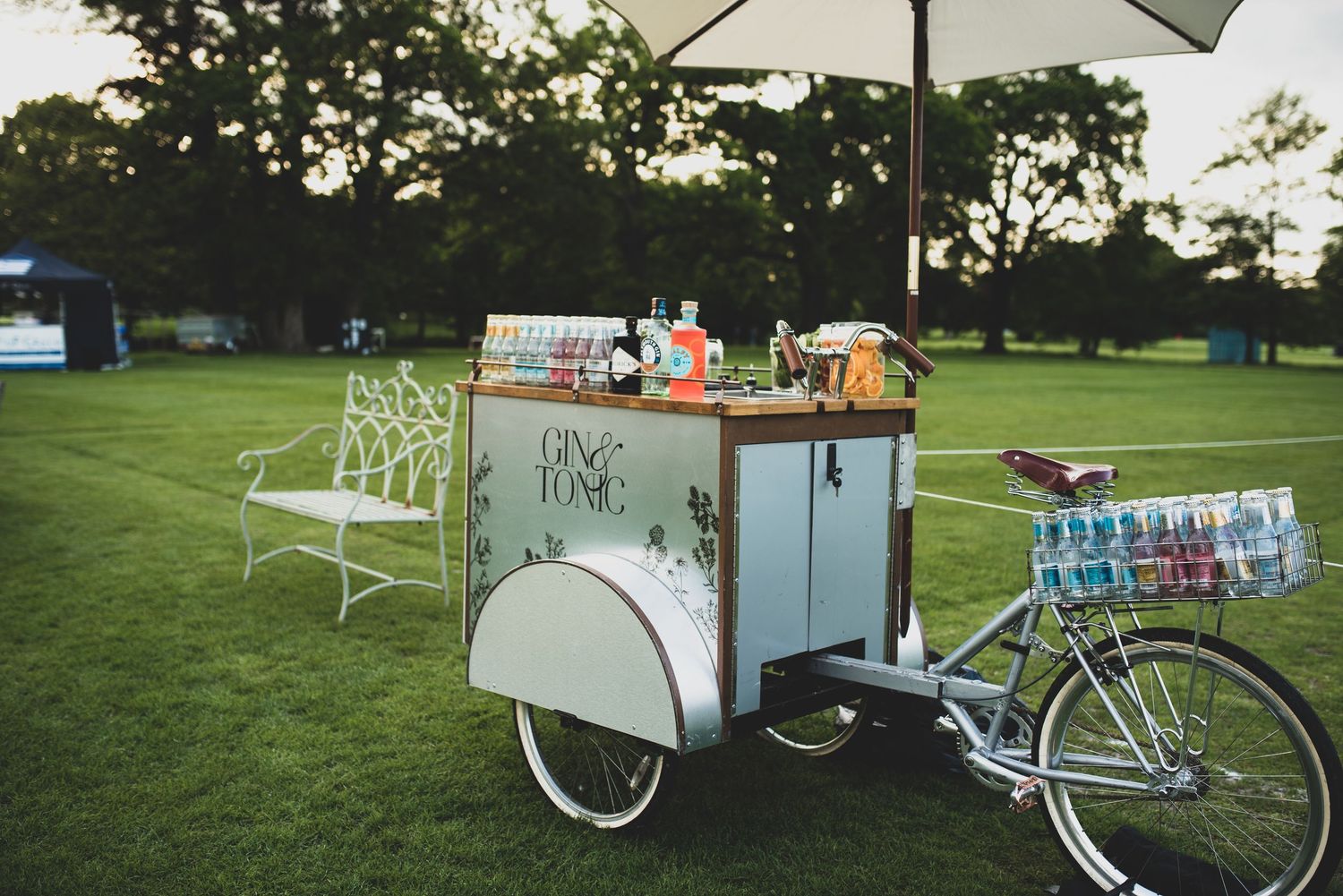 Retro beverage bicycle cart with white umbrella and colorful drinks at dusk outdoor event.
