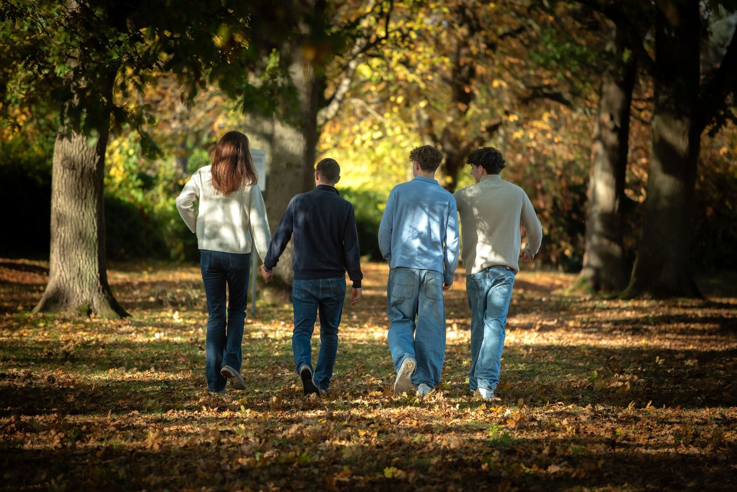 A family of four walks away through a tree-lined park covered in autumn leaves.