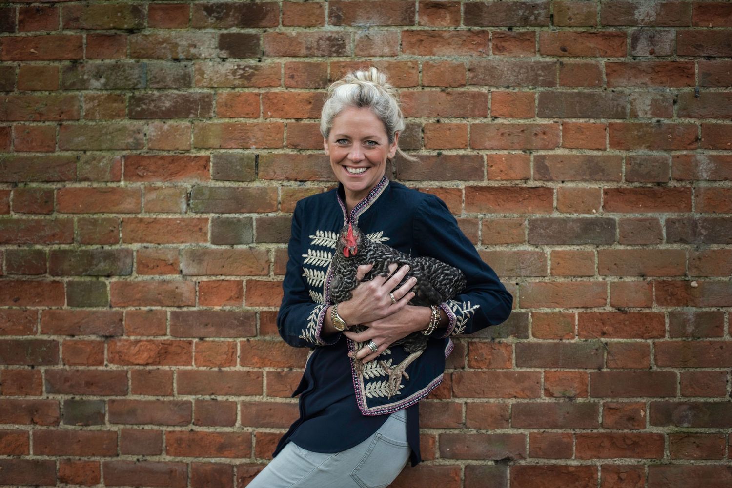 Smiling woman in embroidered jacket holding a black and white chicken against a red brick wall.