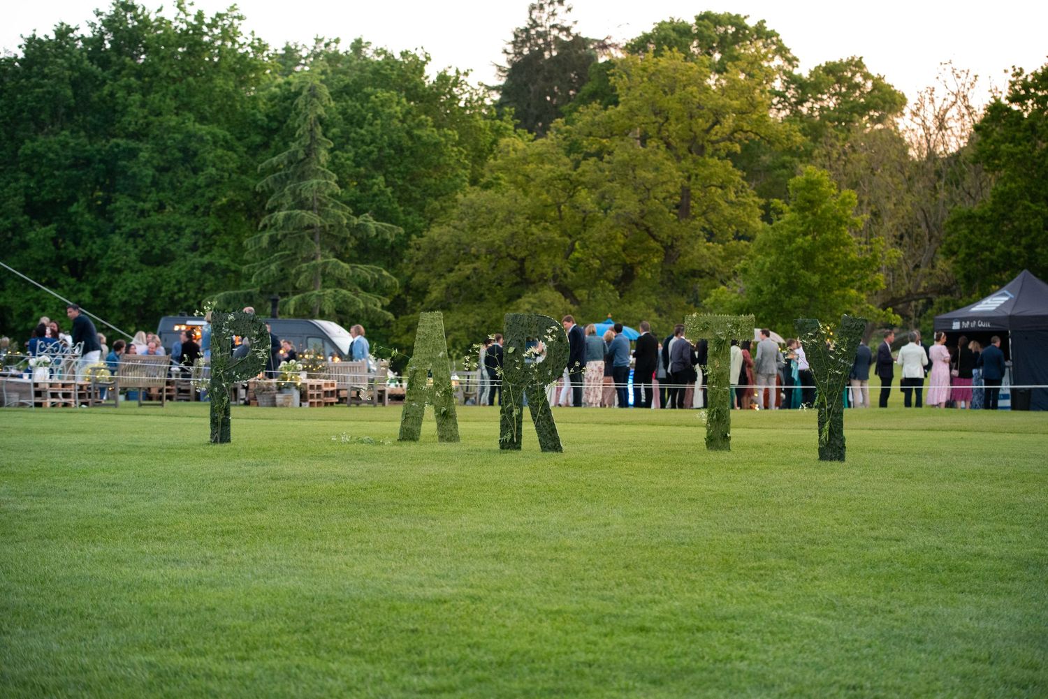 Guests gather on a lush green lawn at an outdoor garden party event at dusk.