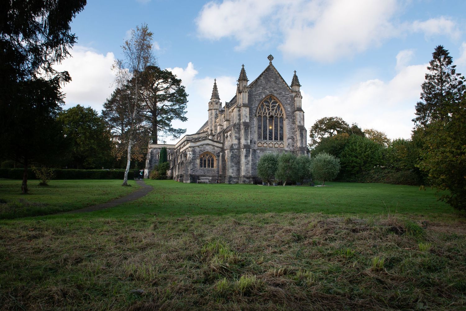 Gothic stone church surrounded by green trees under a partly cloudy sky.  New Forest Wedding. St Saviours Church, Brockenhurst Wedding.