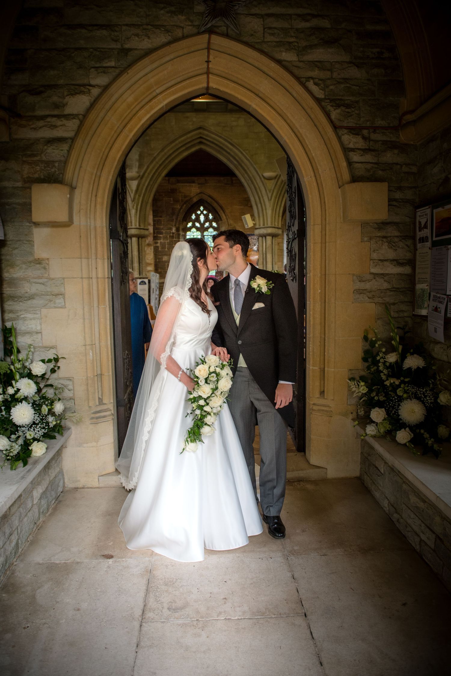 Bride and groom sharing a kiss in a Gothic stone church doorway, bride holding a white floral bouquet. St Saviours Church, Brockenhurst