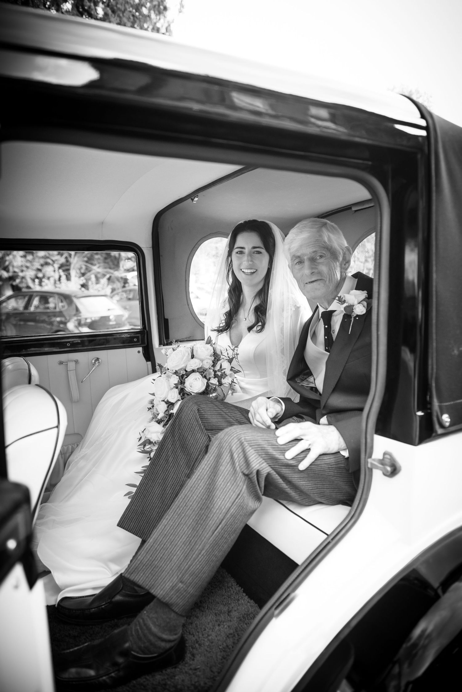 Black and white photo of a smiling bride and father seated in a vintage wedding car.