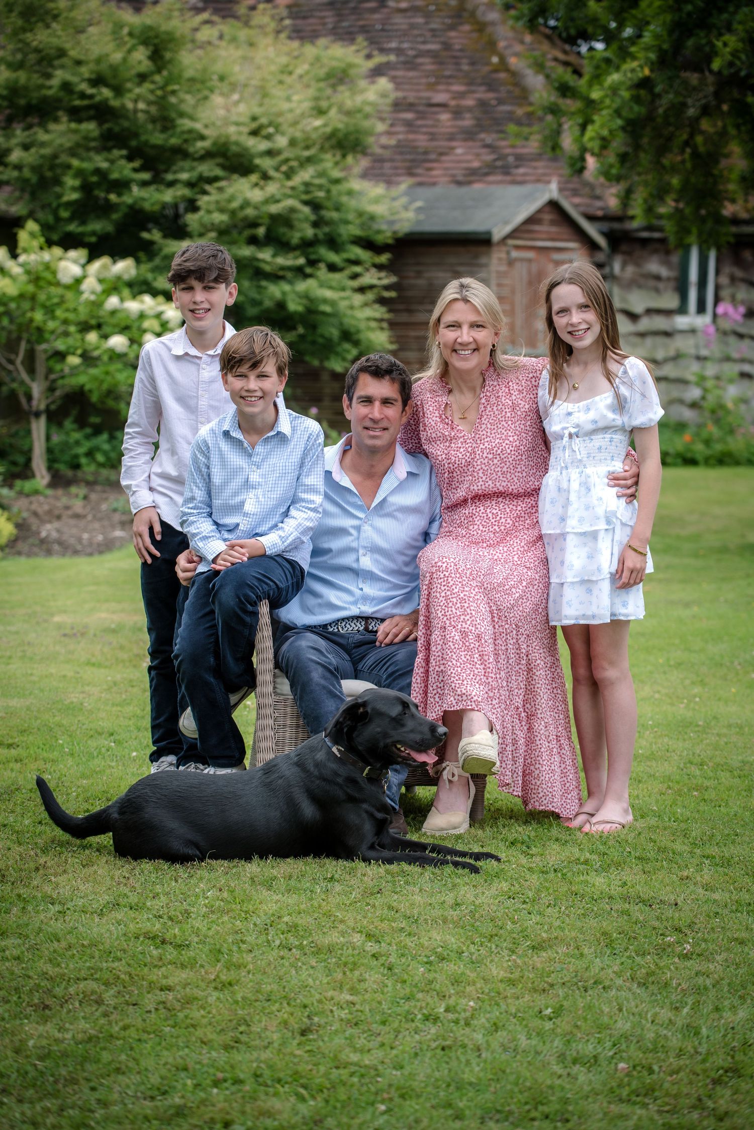 Family of five with black Labrador posing in a lush garden setting.