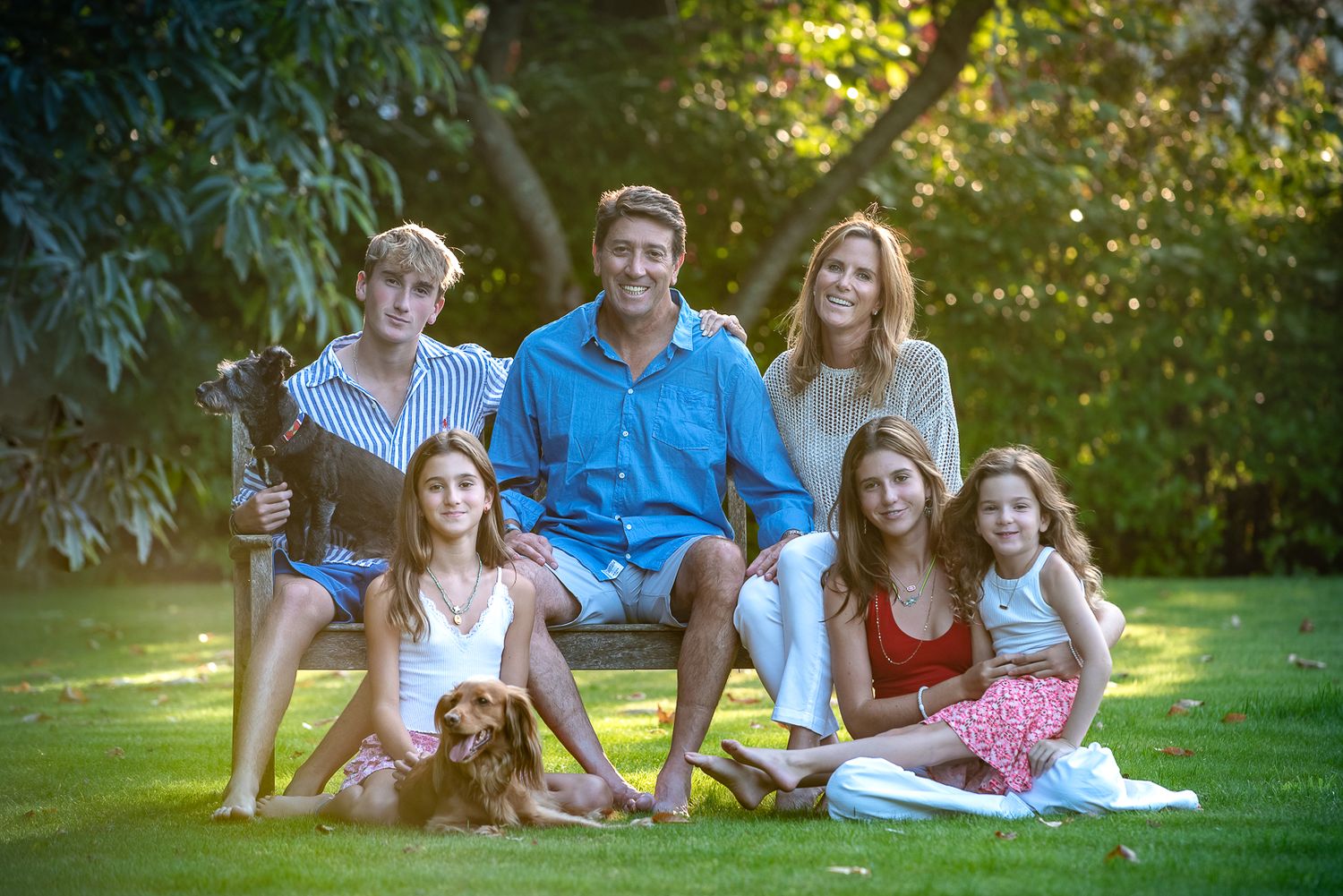 Family of six with dog posing on lawn surrounded by lush green trees during golden hour outdoor photo session.