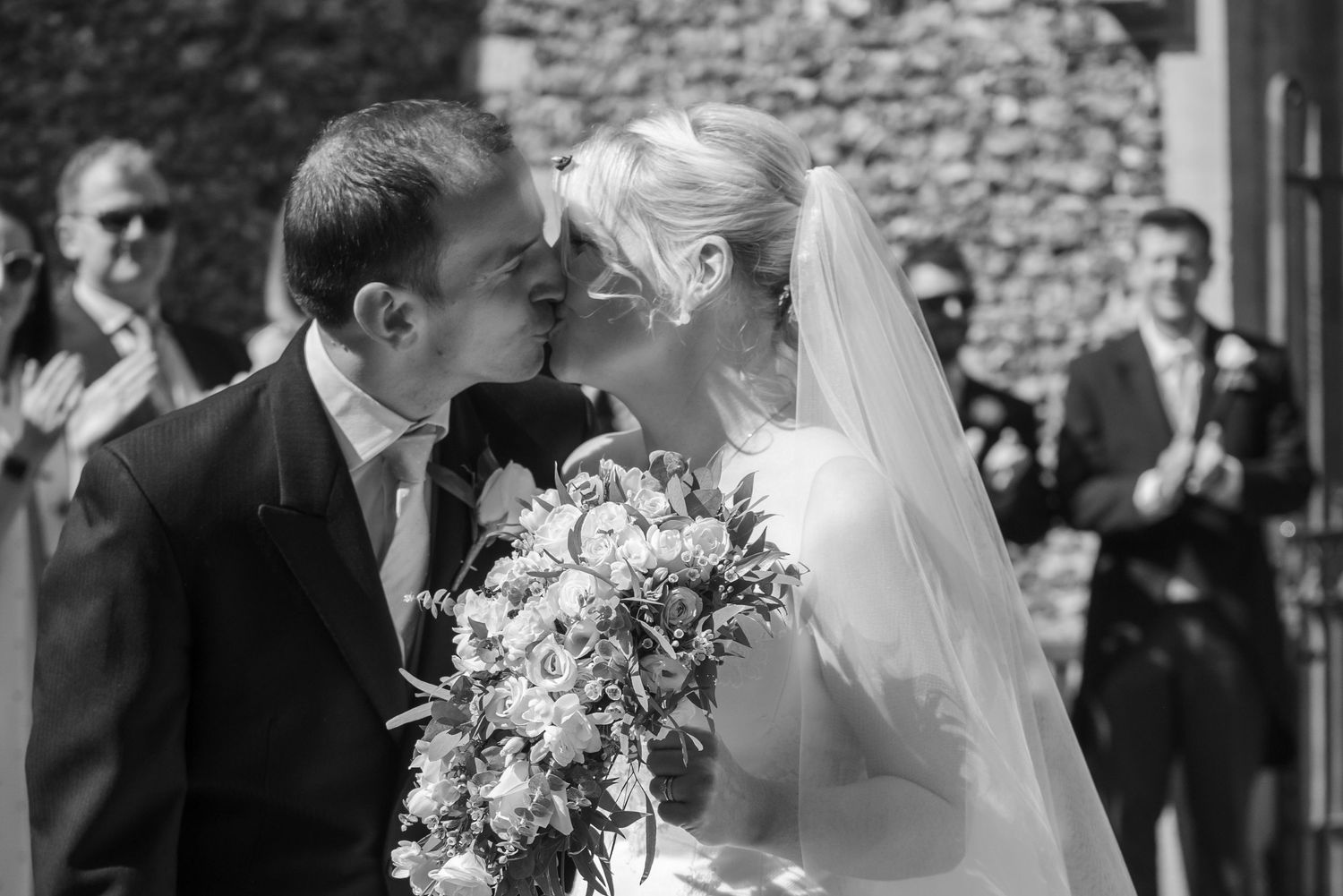 Black and white photo of newlyweds sharing first kiss with wedding bouquet outdoors. St Michaels Church, Bray