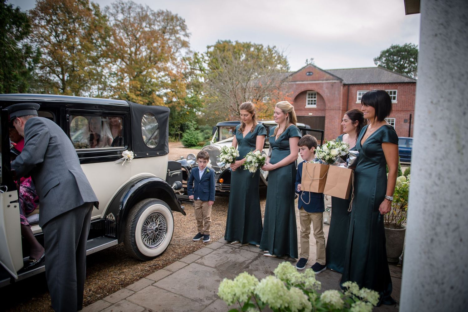 Bridesmaids in teal dresses with bouquets waiting beside a vintage wedding car outside.