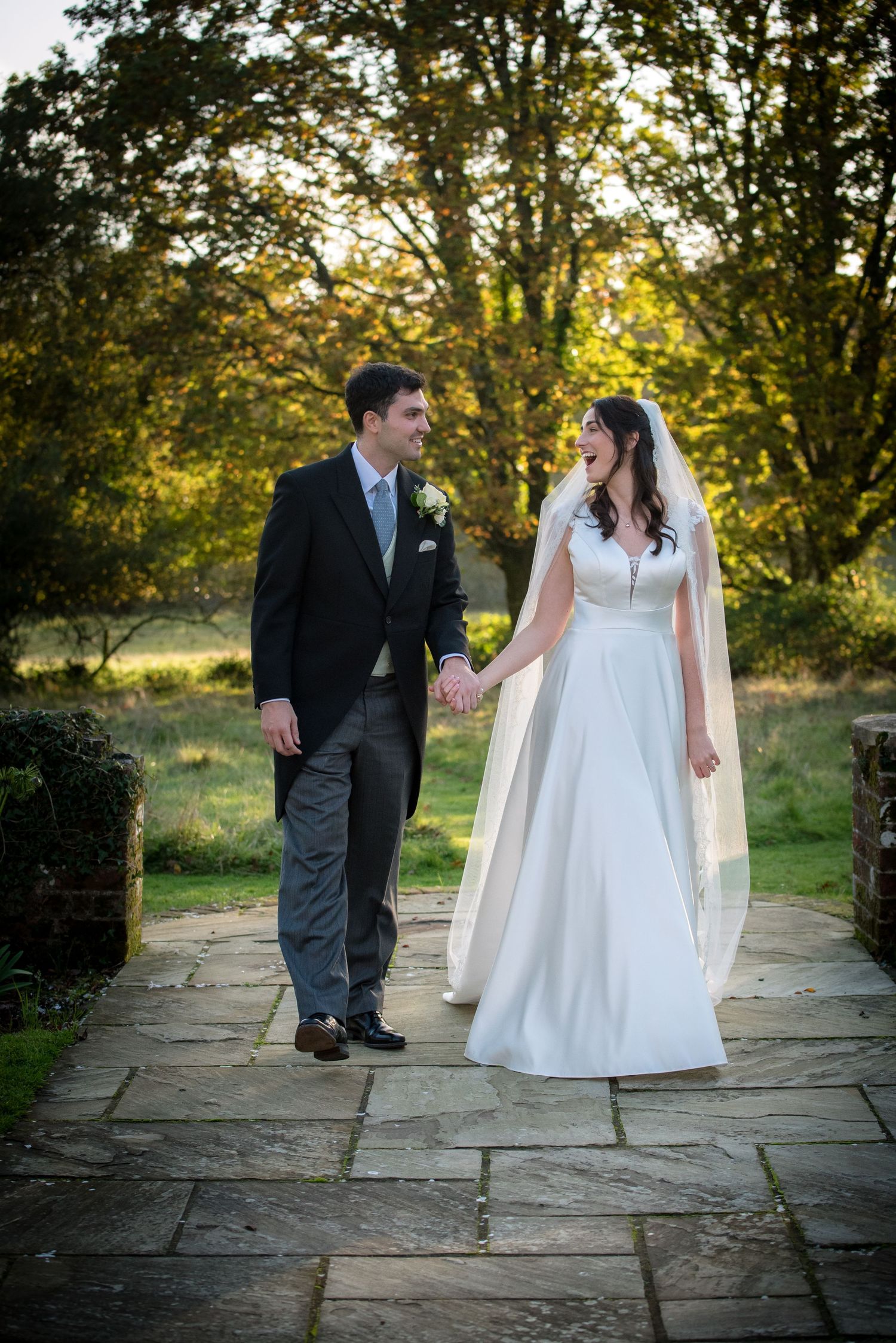 Bride and groom walking hand in hand on a stone path surrounded by golden autumn trees.