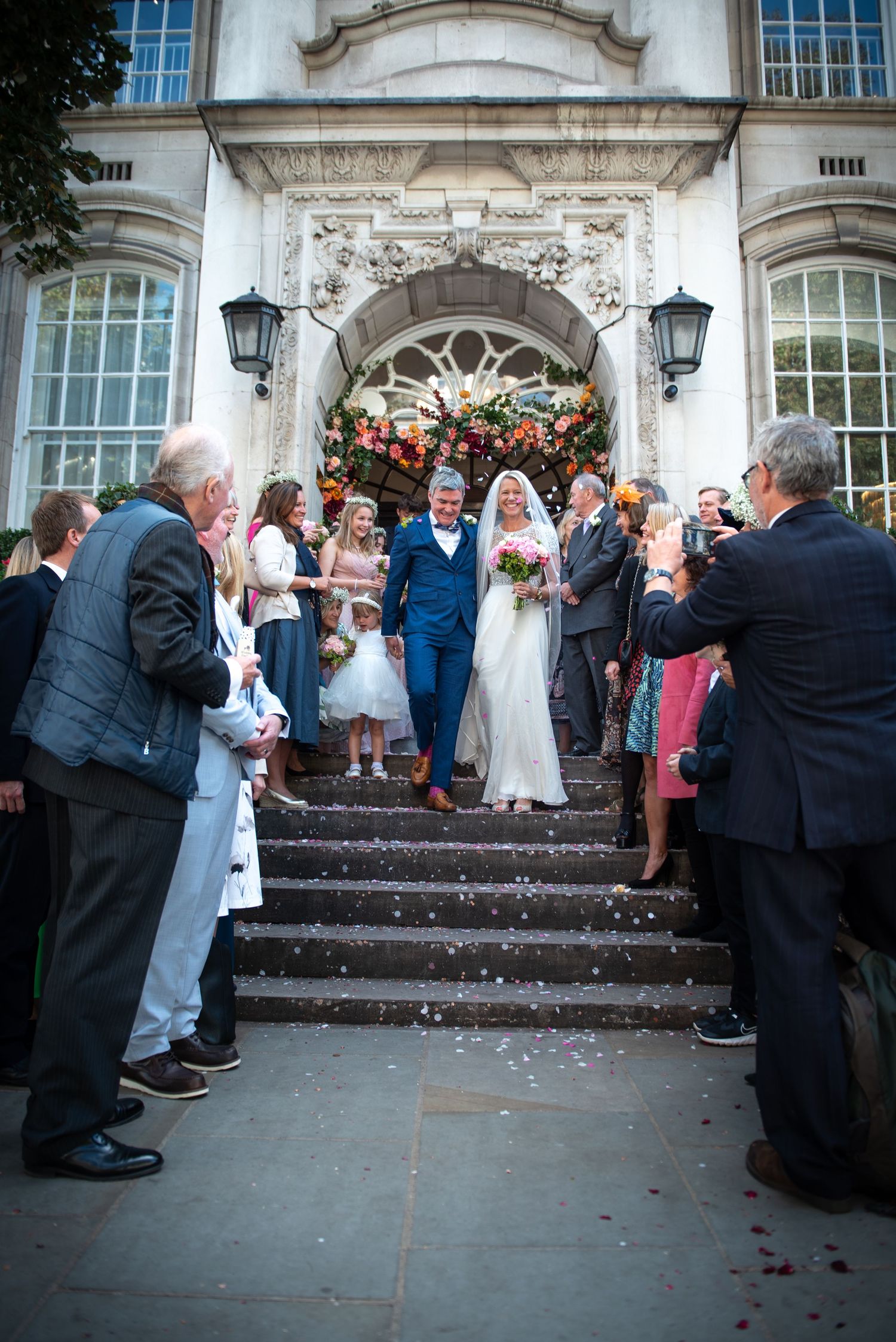 Bride and groom walk down steps through confetti as wedding guests celebrate outside venue.  London Wedding at Chelsea Town Hall.