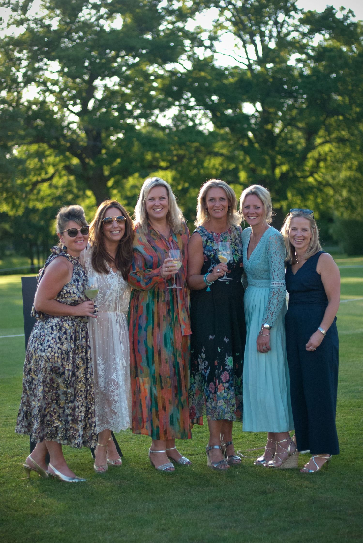 Six women in elegant summer dresses posing together outdoors at a wedding reception on a sunny evening.