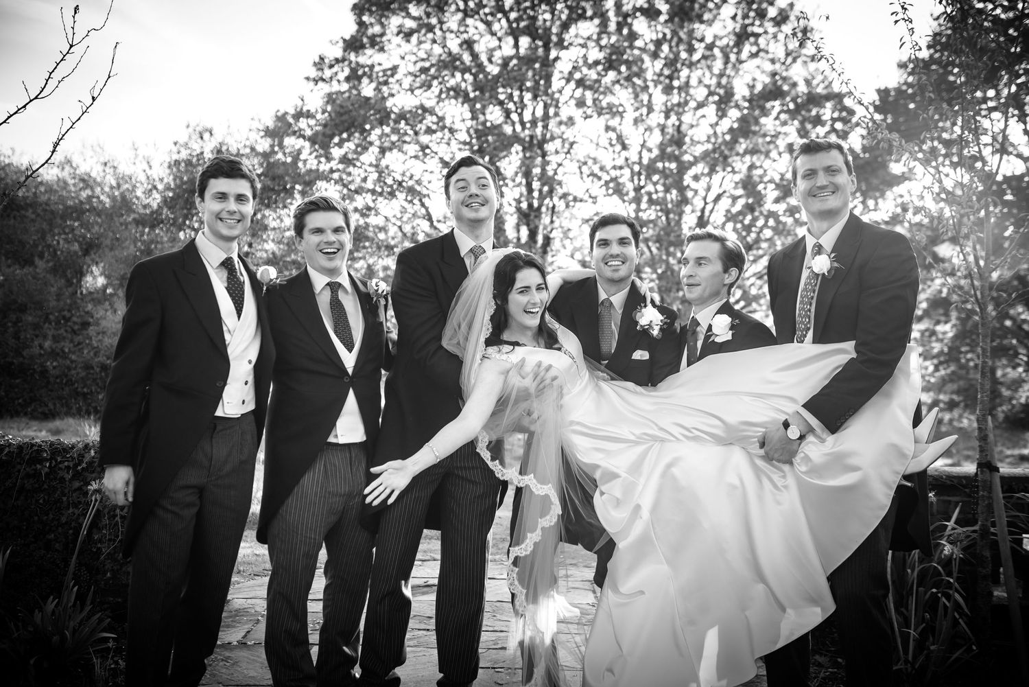Black and white photo of groom being lifted by groomsmen with bride laughing outdoors.