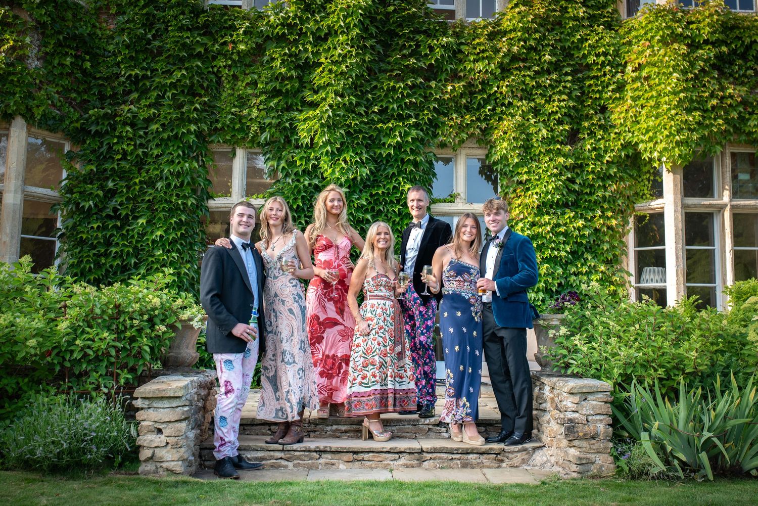 Seven smartly dressed guests smile together on stone steps outside a lush ivy-covered estate. event photography