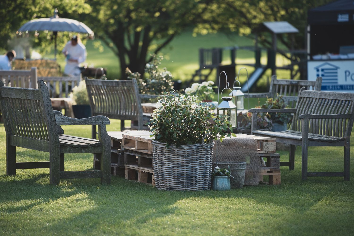 Rustic outdoor garden lounge with wooden chairs, wicker basket planter, and lush green lawn. Event set up