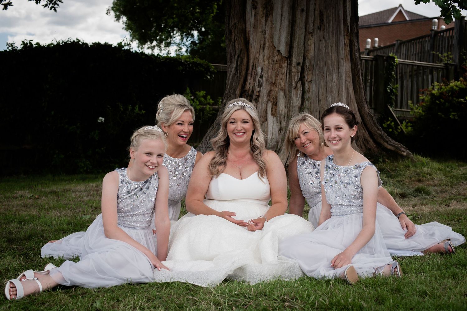 Bride in white gown sits with four bridesmaids in silver dresses on grass by large tree.