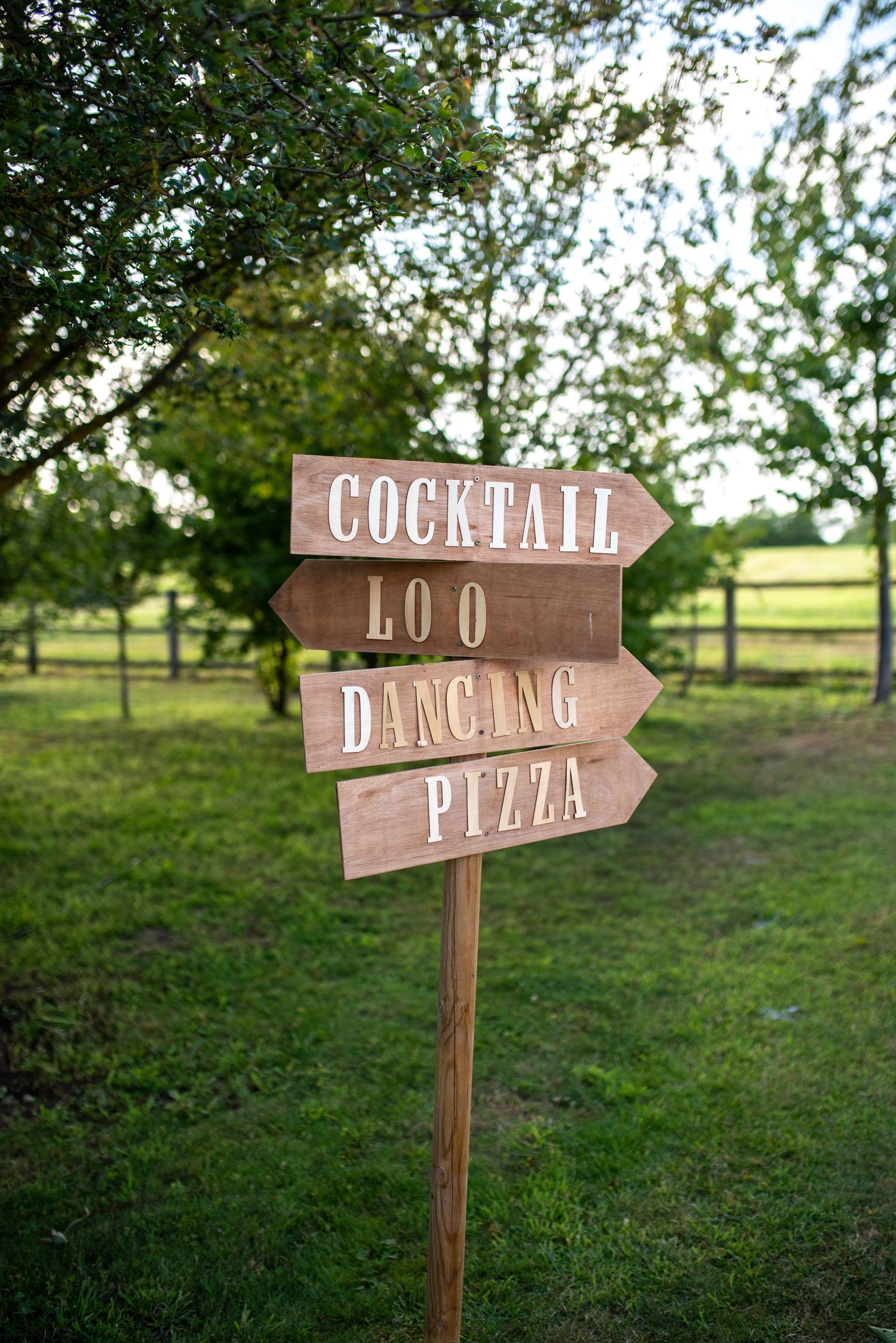 Rustic wooden directional sign pointing to cocktail, loo, dancing, and pizza at an outdoor event.  events party