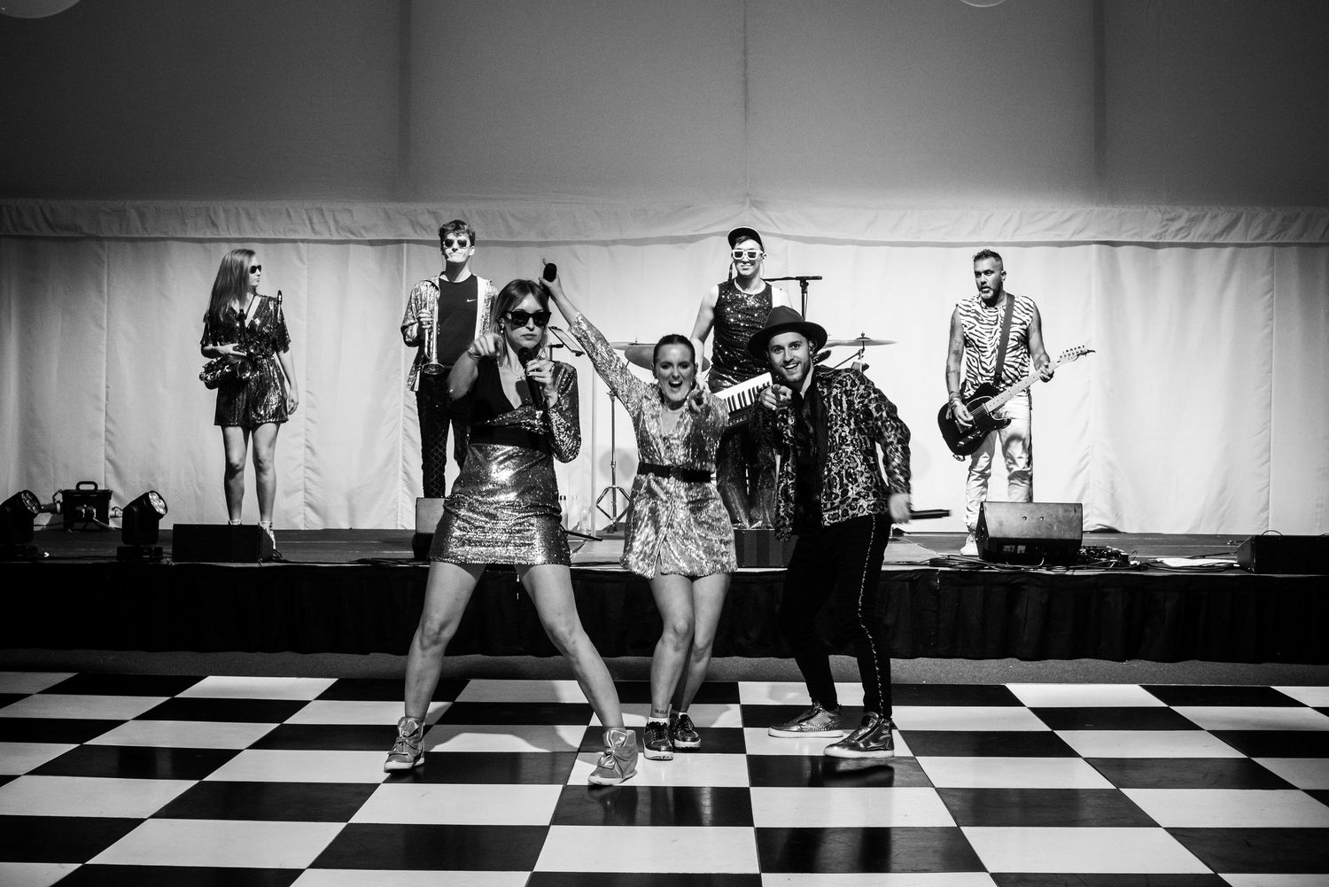 Black and white photo of energetic band performing on checkered dance floor in marquee venue. Event photography