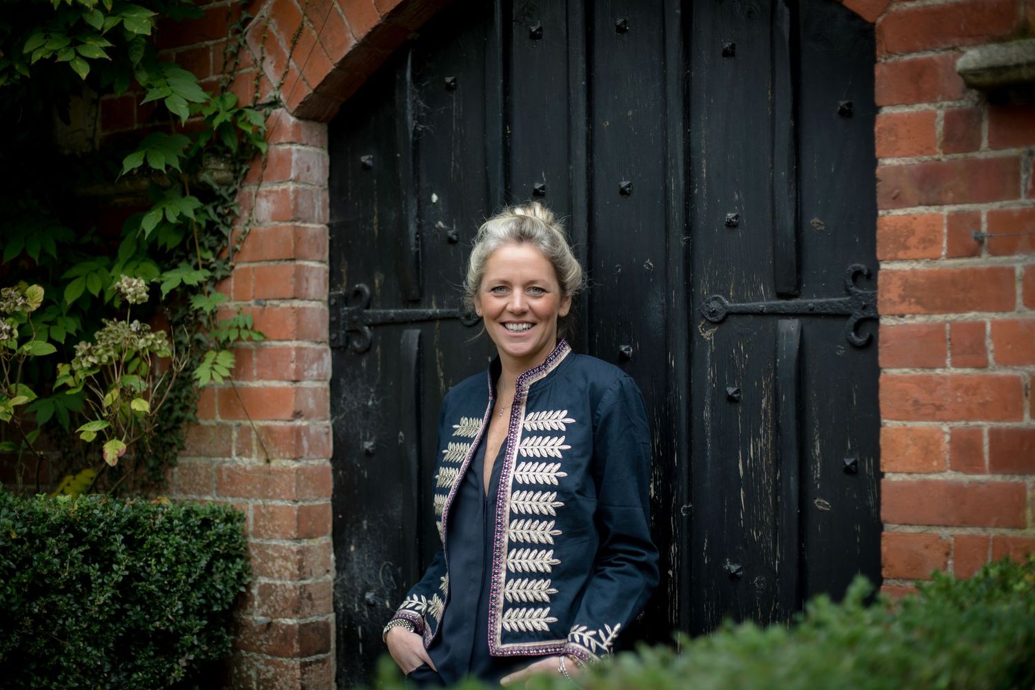 Smiling blonde woman in embroidered navy jacket standing before charming black wooden doors. business portrait