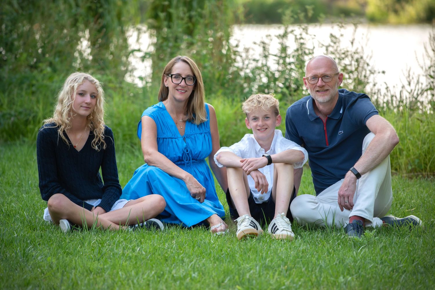 A family of four sits together on green grass near a peaceful lakeside setting.