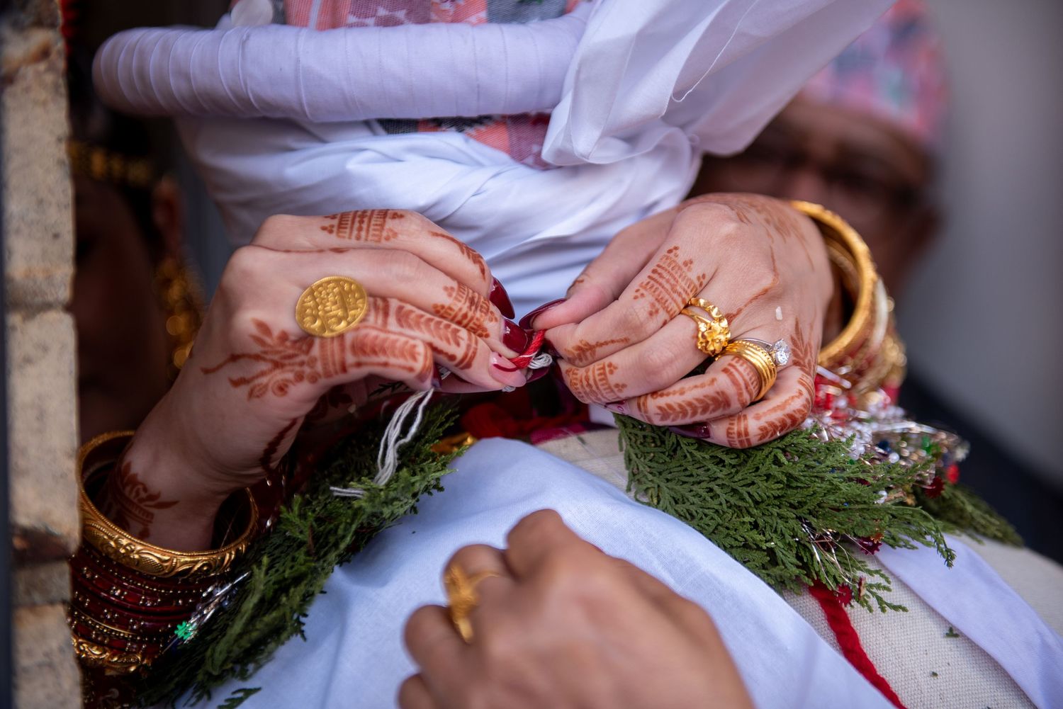 Close-up of bride and groom hands adorned with henna, gold rings, and bangles holding greenery.