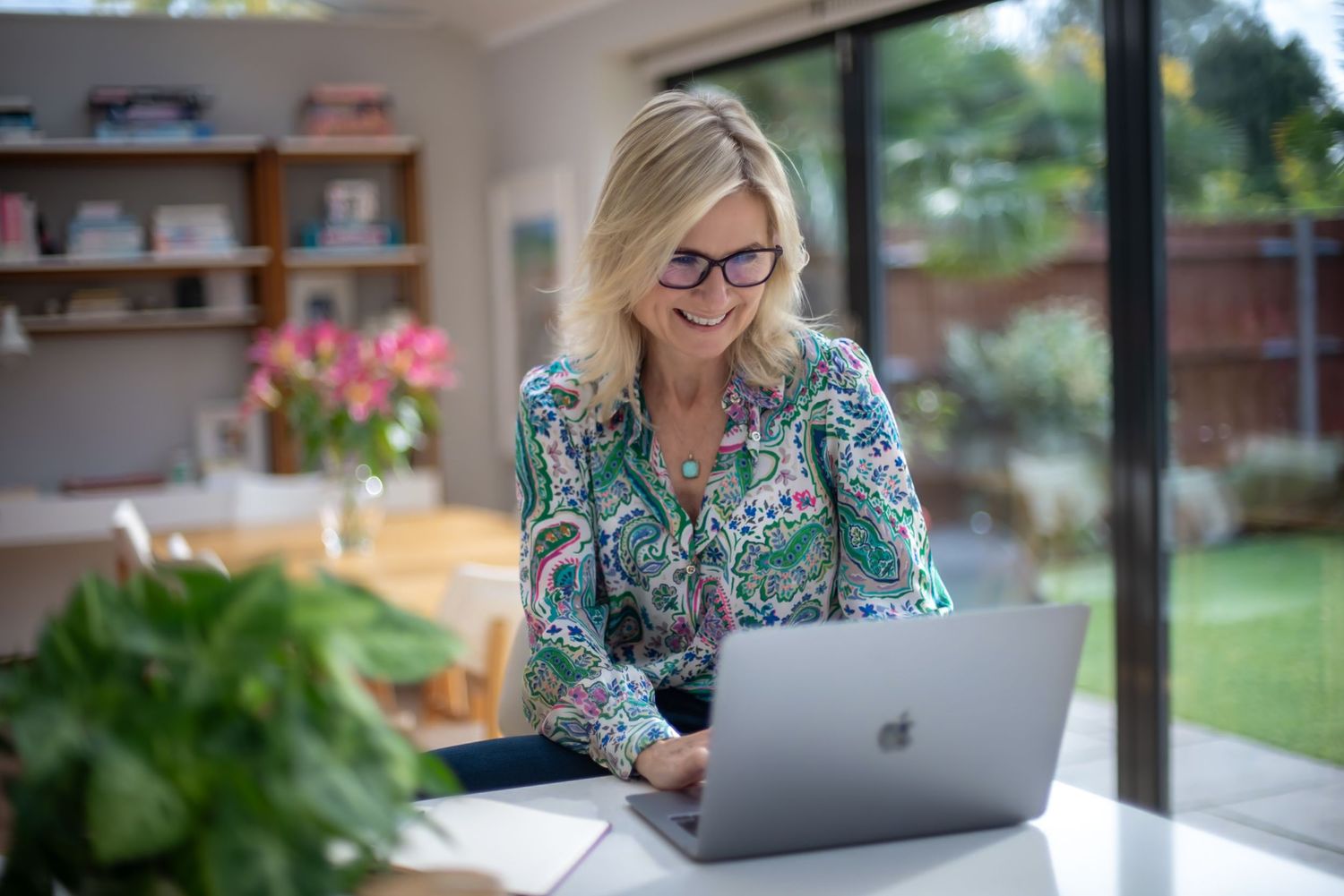 Blonde woman wearing glasses smiling while working on a laptop at a bright home office desk. business portrait