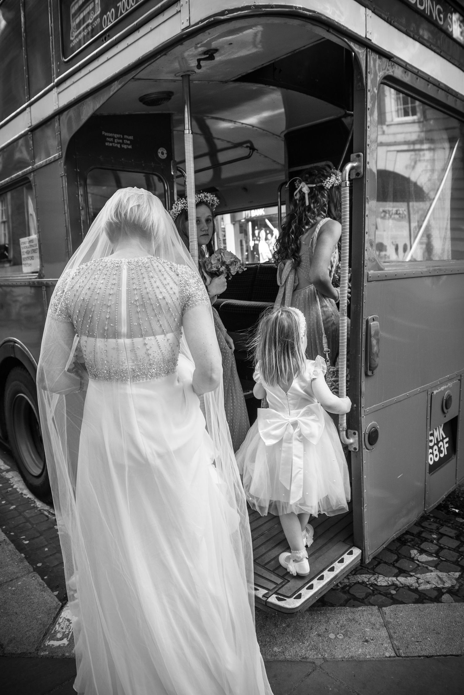 Bride in white gown and veil with flower girl boarding a red double-decker bus on wedding day.