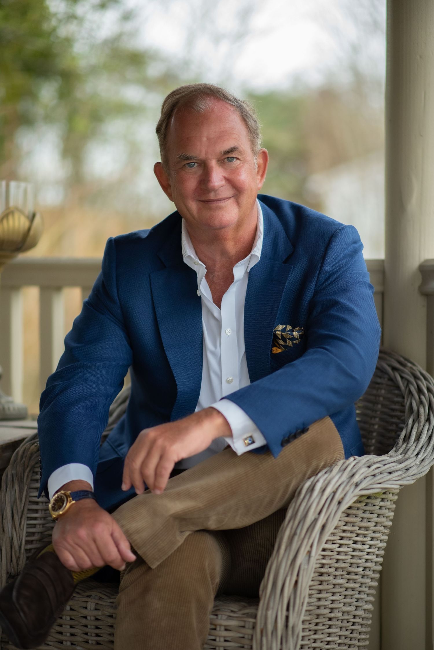 Distinguished man in blue blazer sitting relaxed in a wicker chair on a porch portrait.  John Spence, Karma Resorts