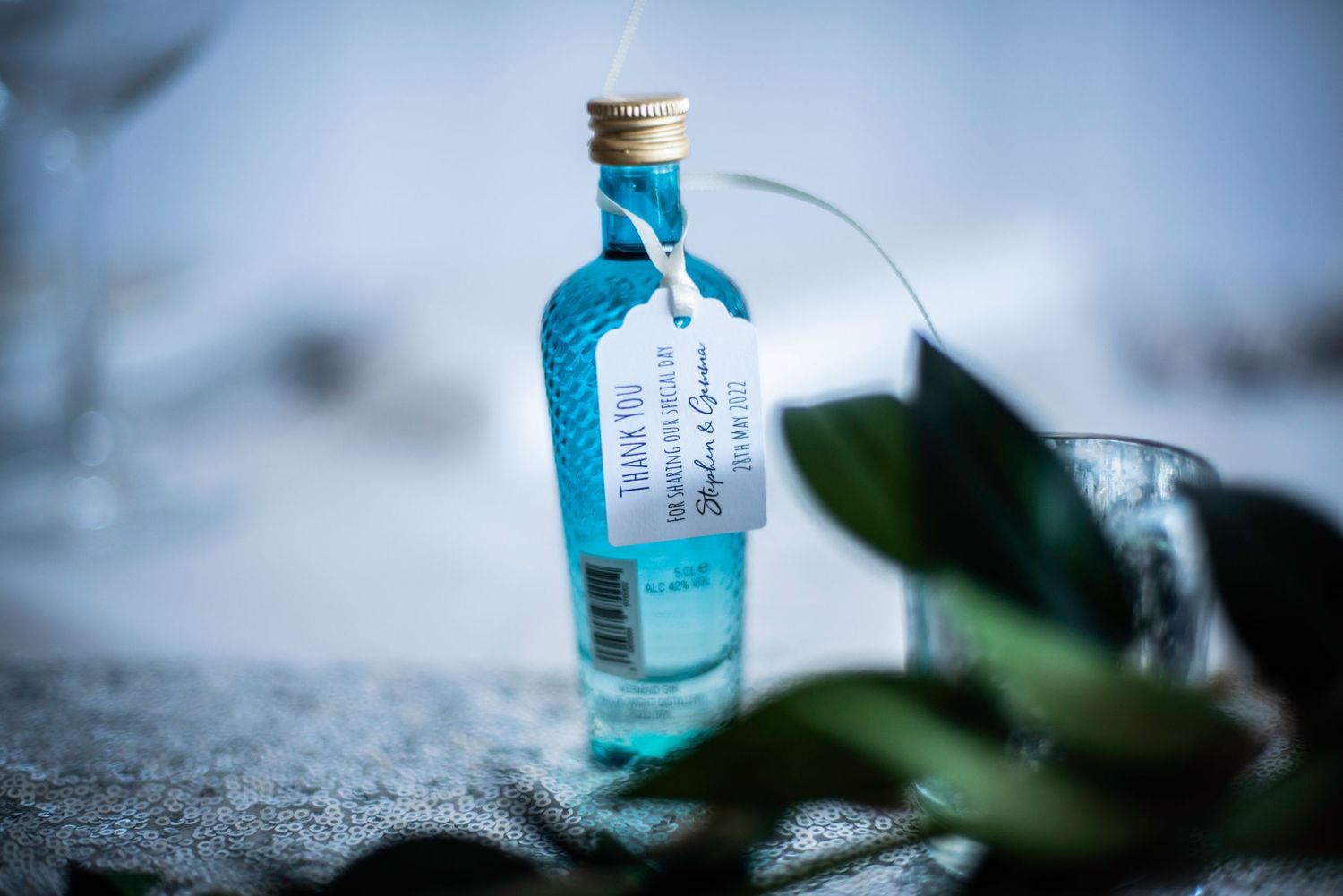 Blue gin bottle wedding favor with personalized tag among green leaves on reception table.