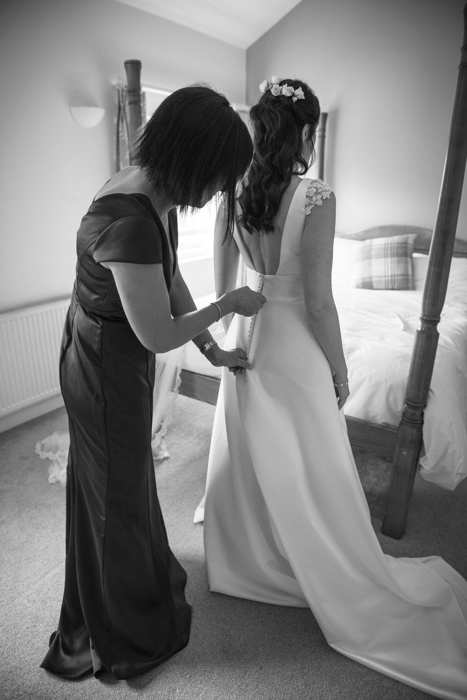 Bridesmaid helping bride with wedding dress buttons in a bedroom, black and white.