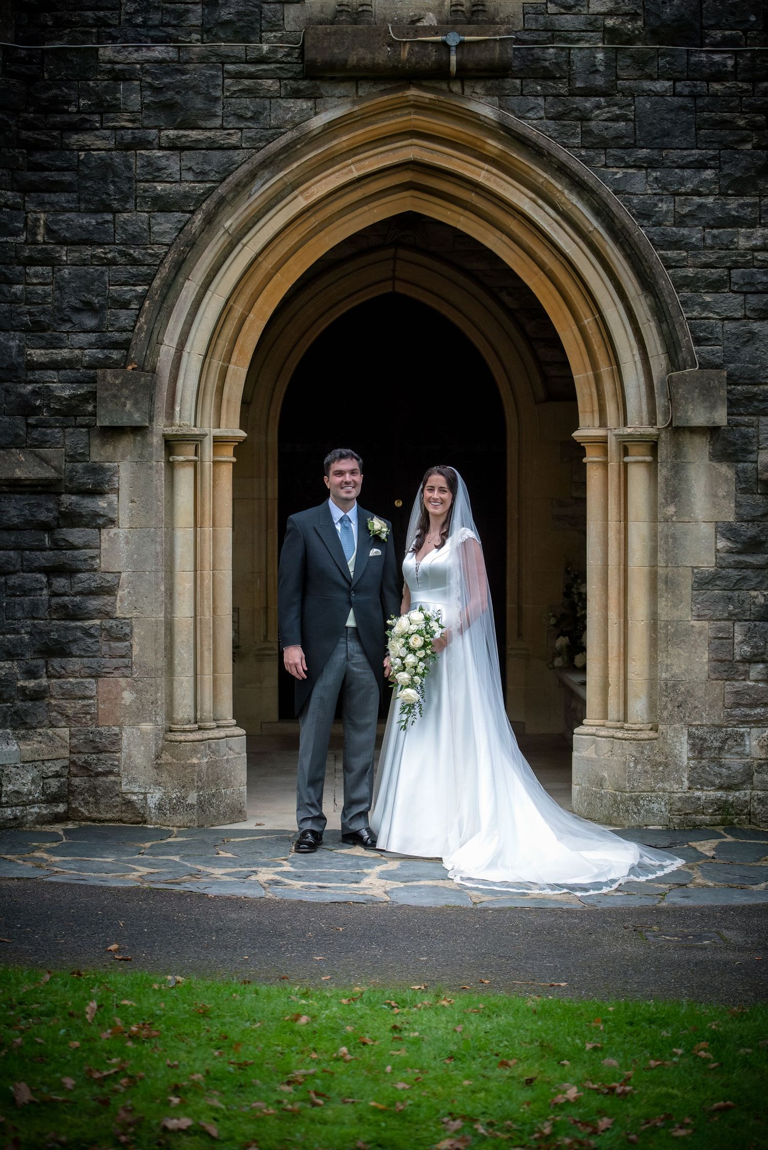 Bride and groom posing together under Gothic stone arch doorway of historic church after wedding ceremony.  St Saviours Church, Brockenhurst